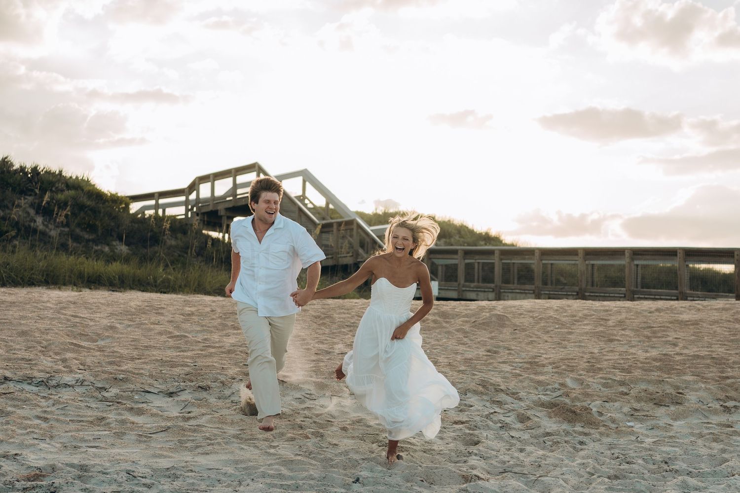 A couple in white clothing runs playfully along the beach at sunset.