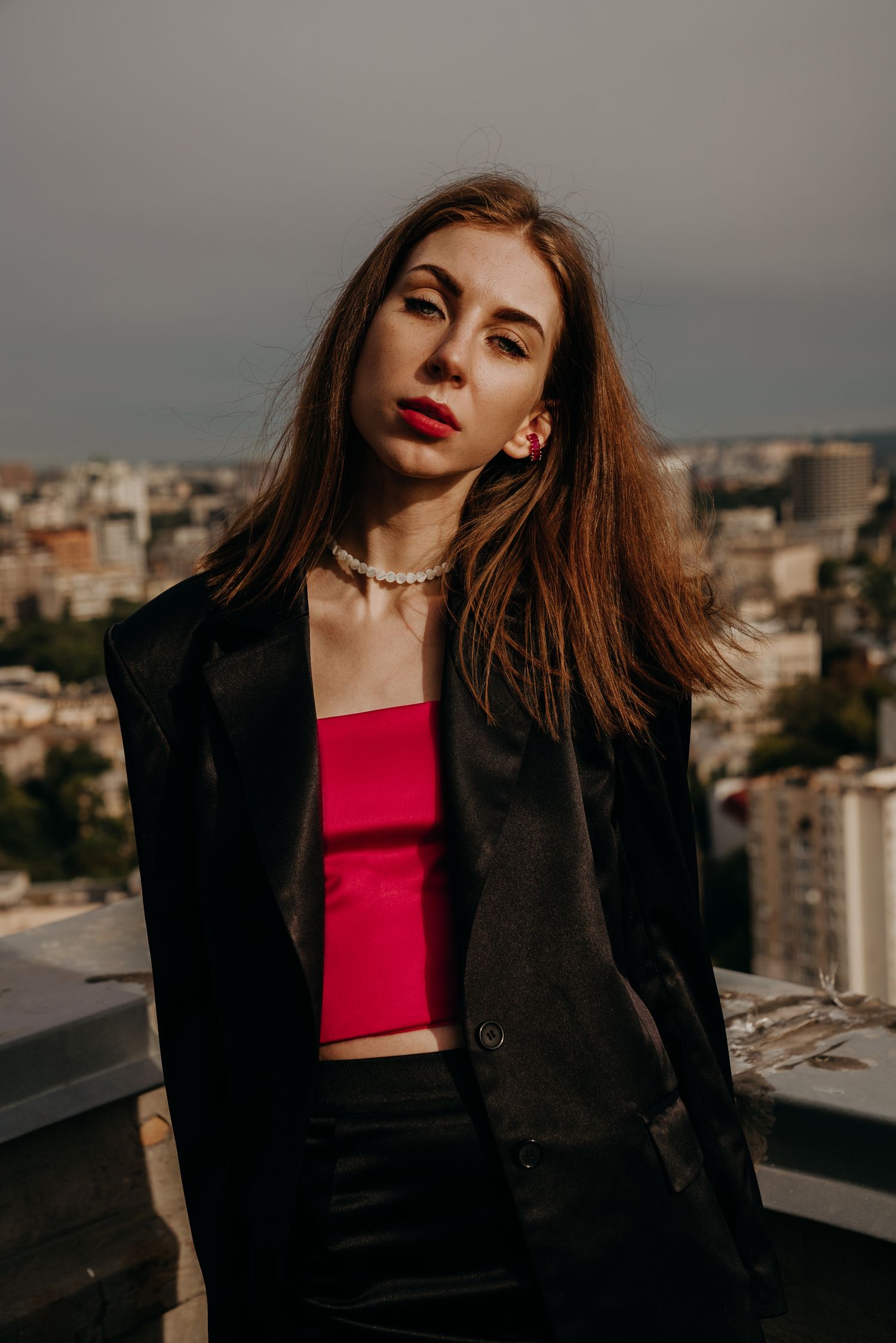 Young woman in black blazer and red crop top with pearl necklace posing on rooftop with city skyline background.