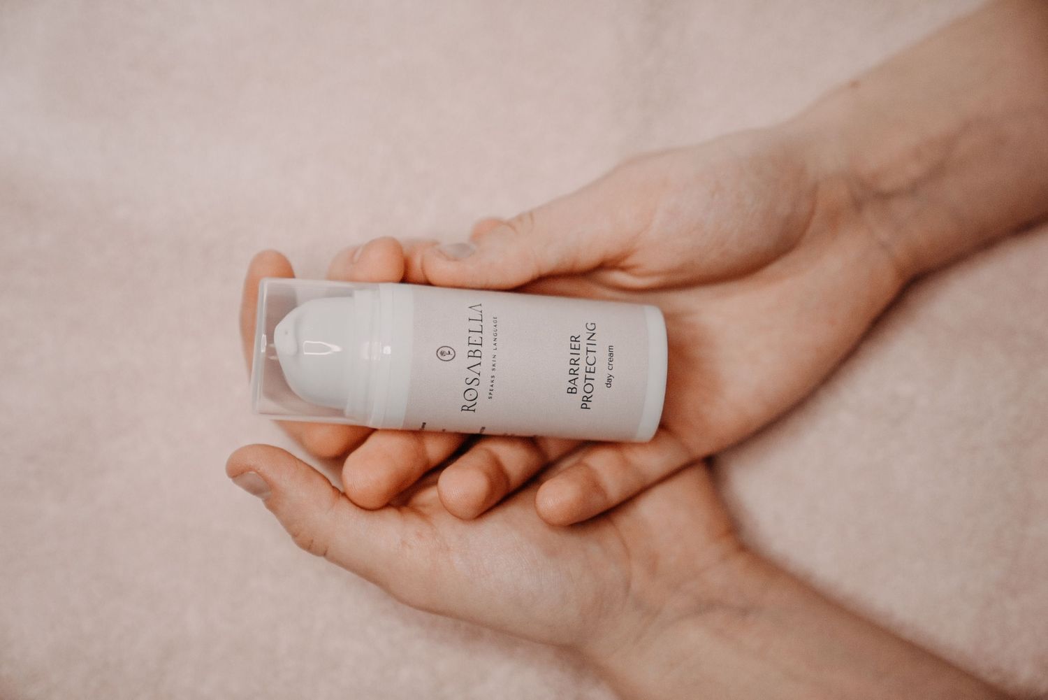 Hands holding a small white Rosabelli skincare serum bottle against a soft pink background.
