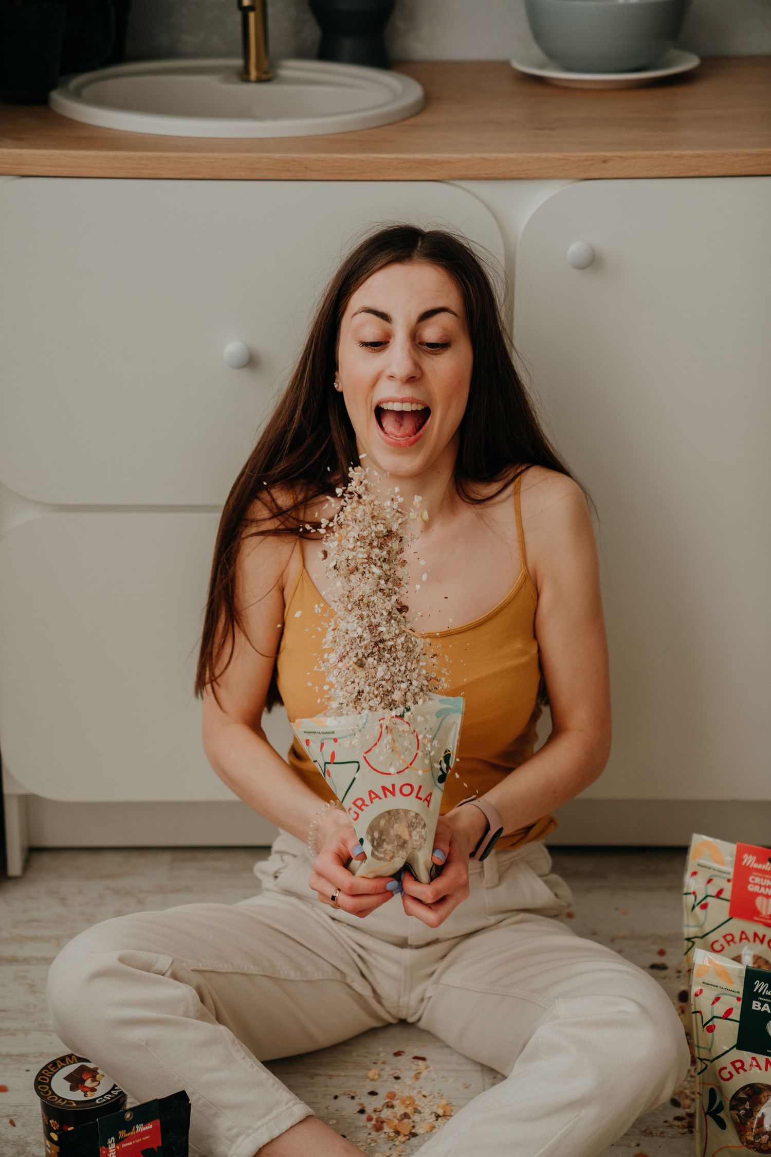 Excited woman sitting on kitchen floor with granola flying from a bag toward her open mouth.