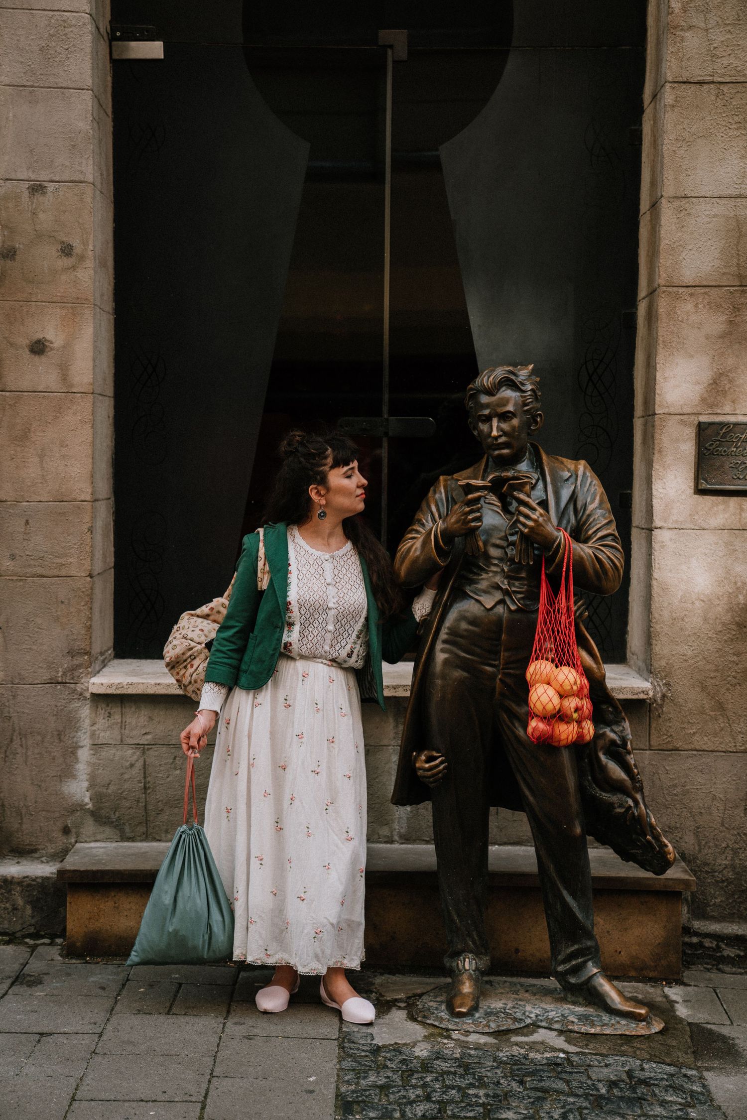 Woman in white dress standing beside bronze statue holding red mesh bag with apples.