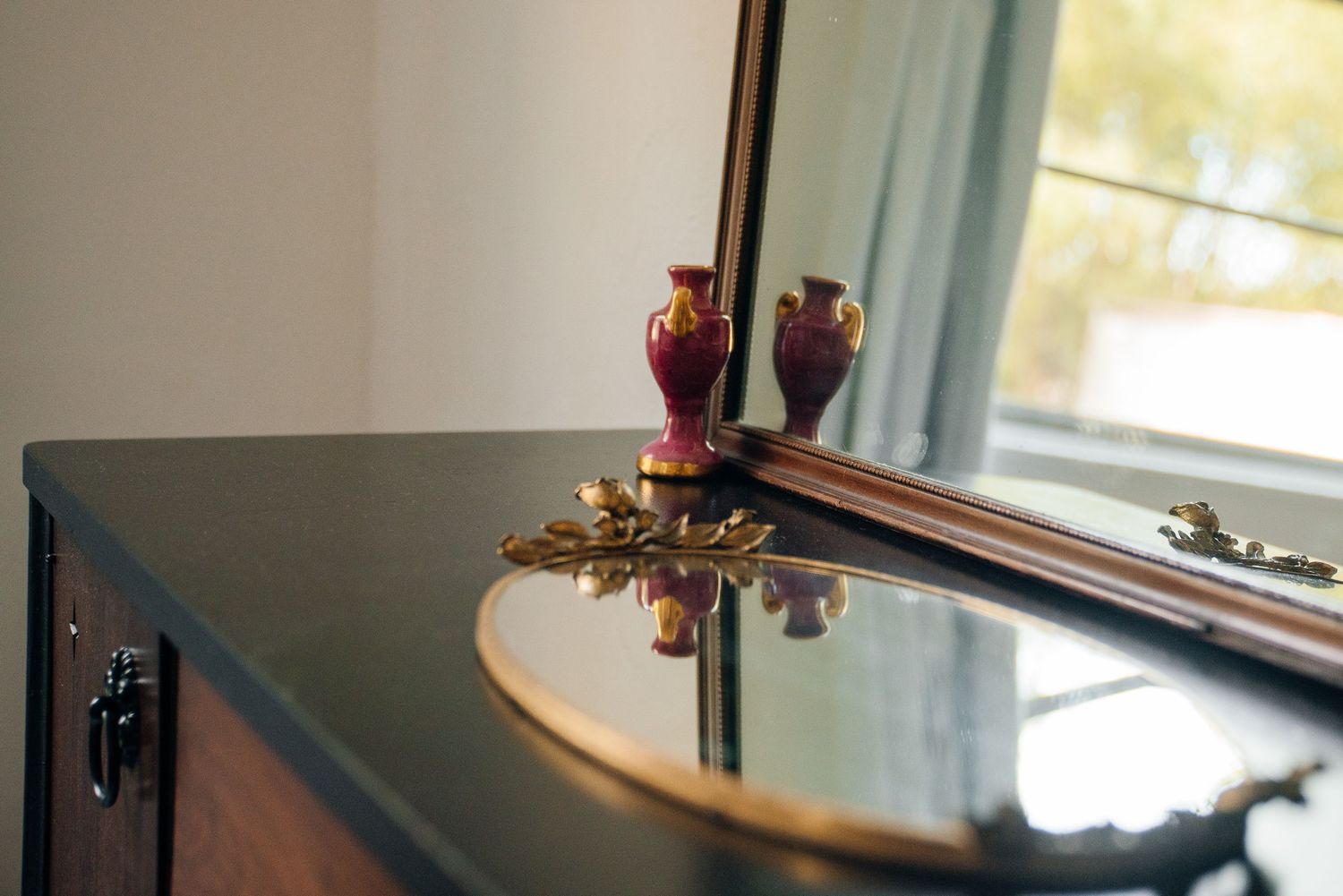 Elegant dark dresser with ornate gold details, two red vases, and mirror reflection near a bright window.
