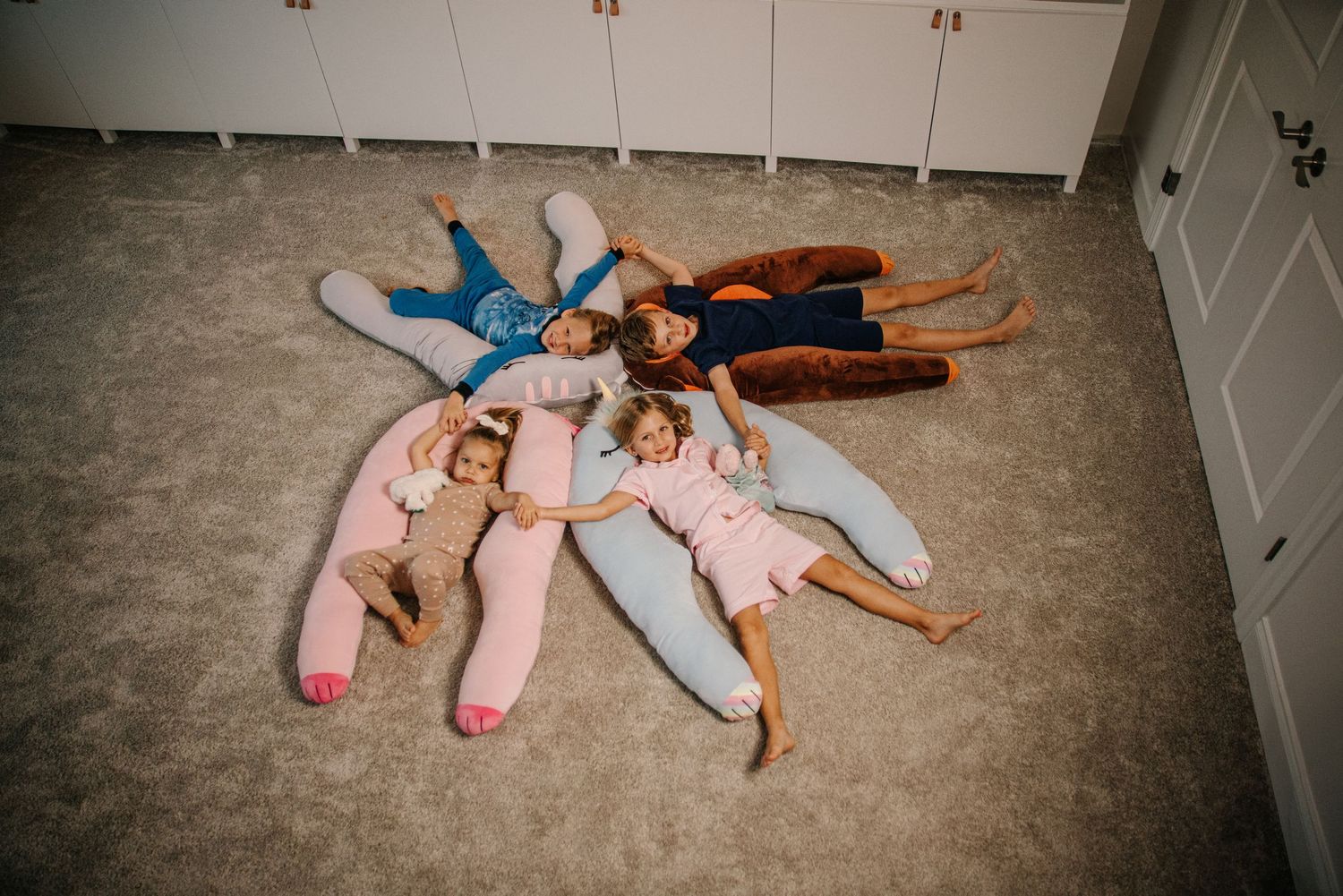 Four children lying on floor in a star pattern with colorful body pillows in pink, blue, and brown.