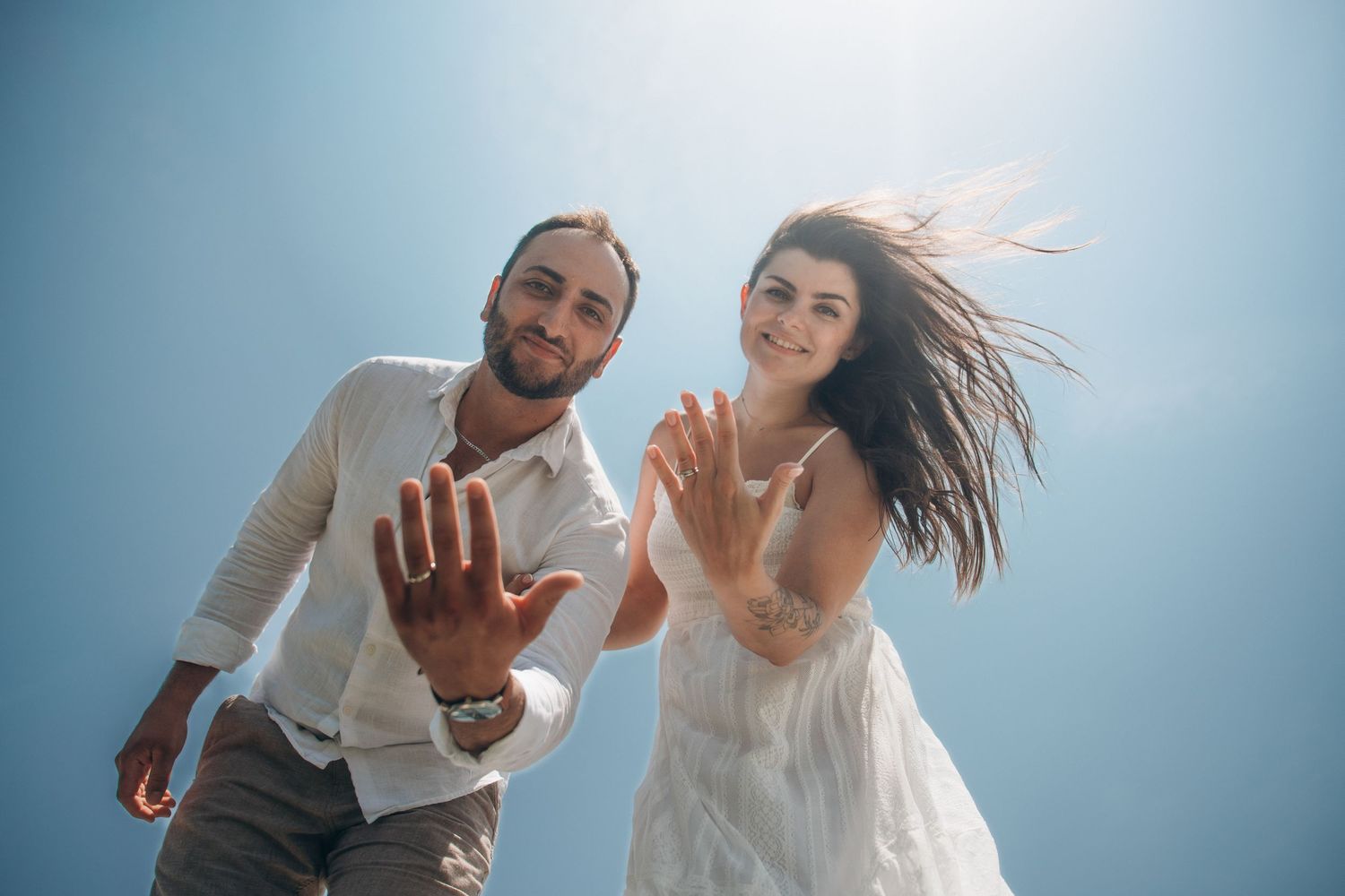 Happy couple in white attire reaching toward camera against blue sky, woman's hair flowing in wind.