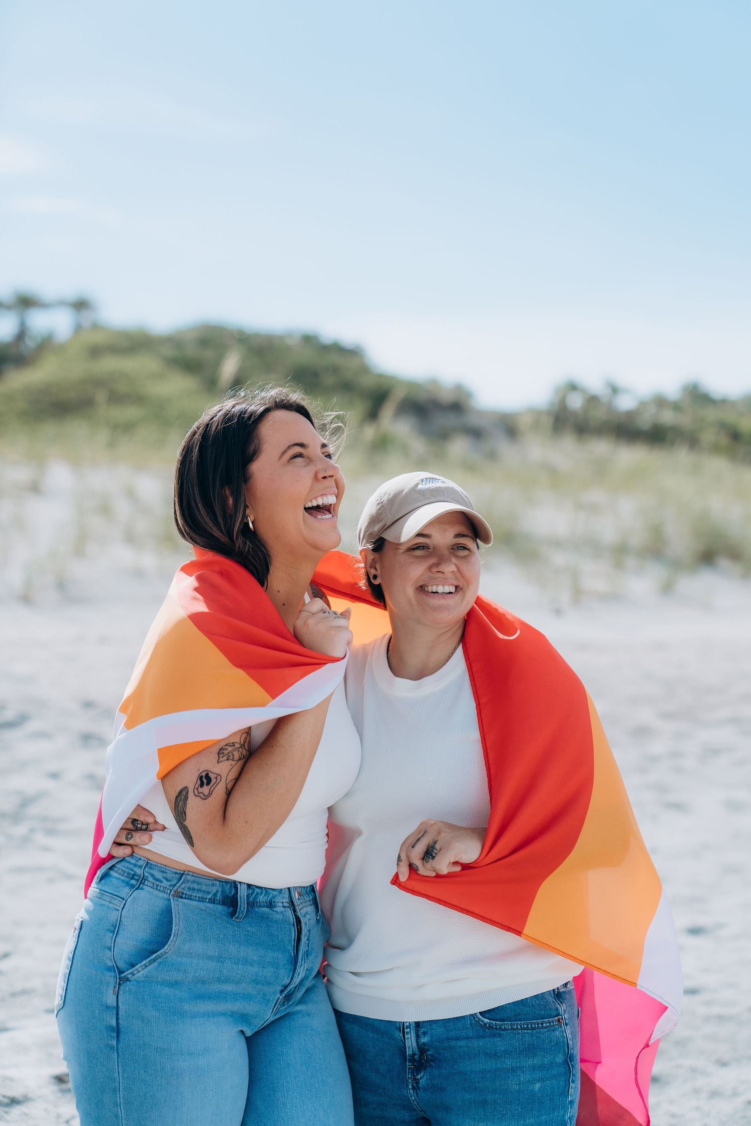 Two people laughing together while wrapped in a vibrant rainbow flag at the beach.
