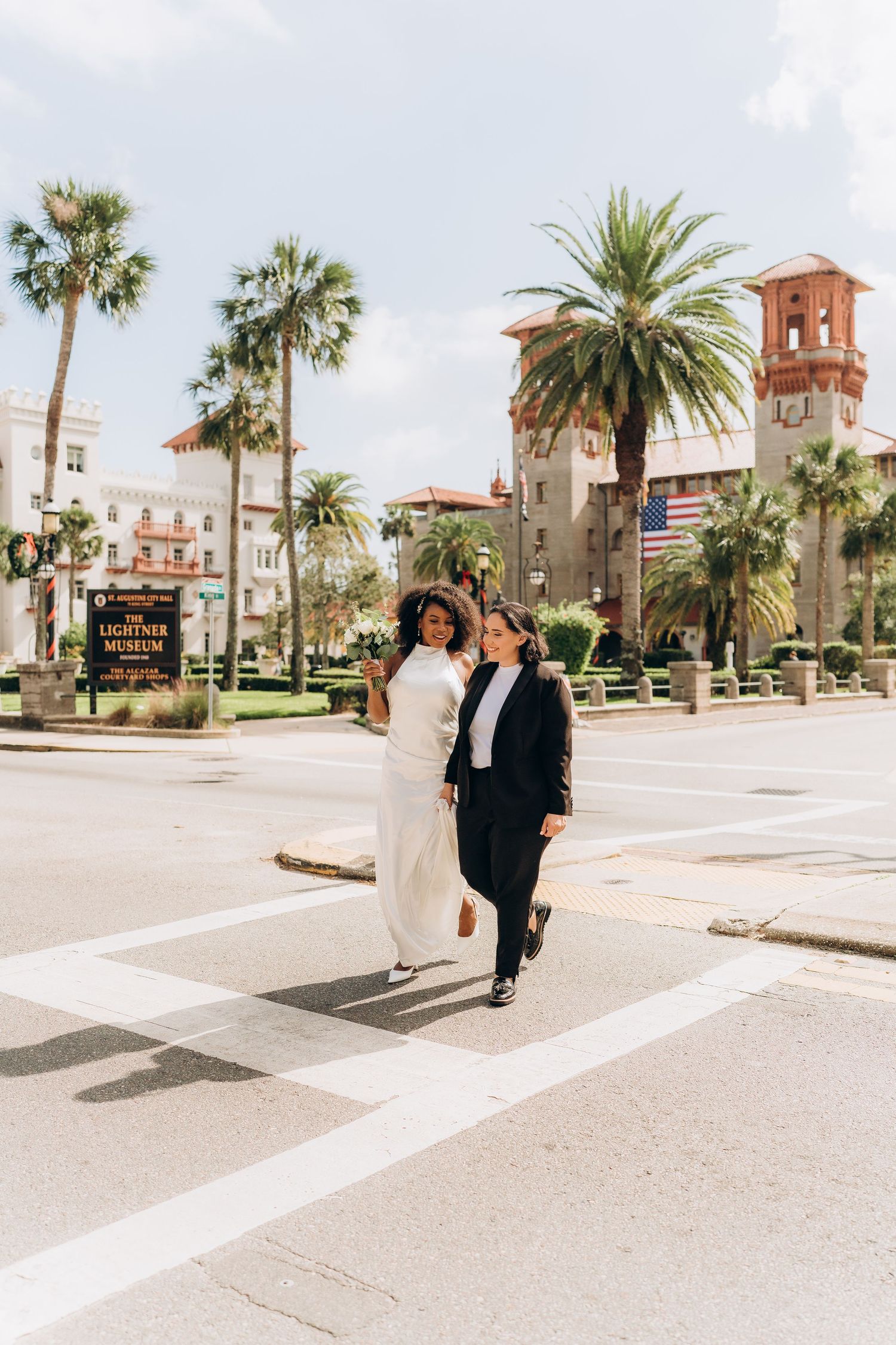 A couple in wedding attire crosses a sunny plaza with palm trees and Spanish architecture in the background.