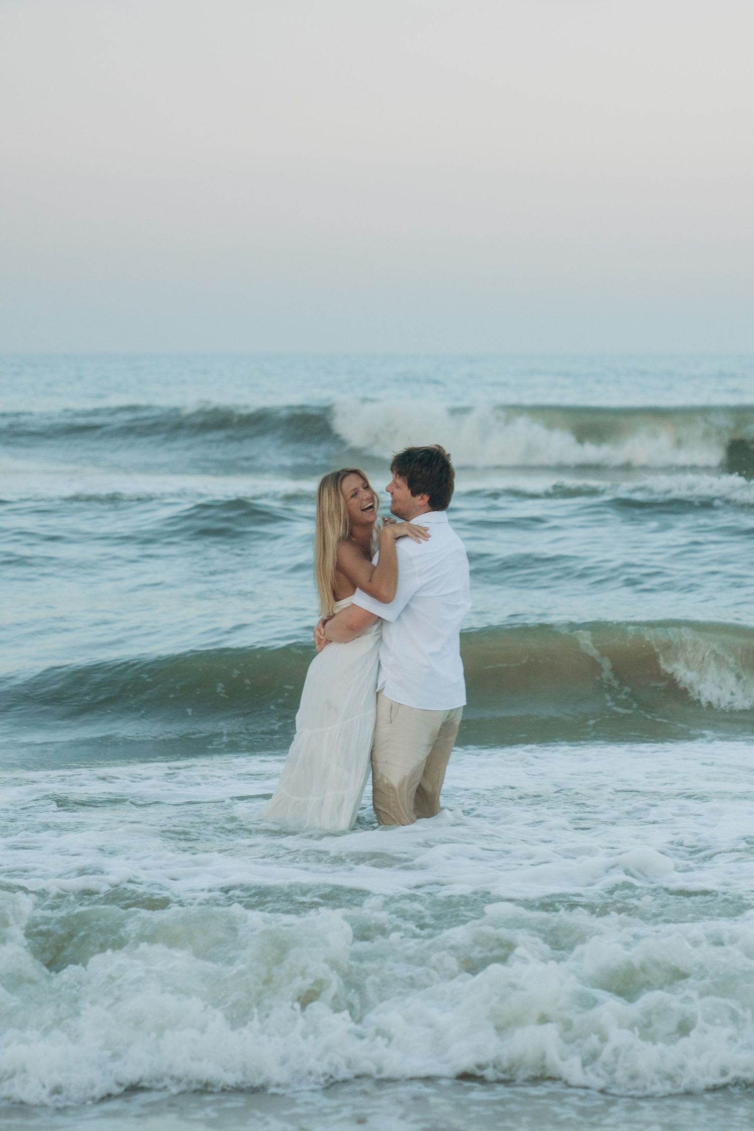A couple in white clothing stands together in the ocean as waves crash around them.
