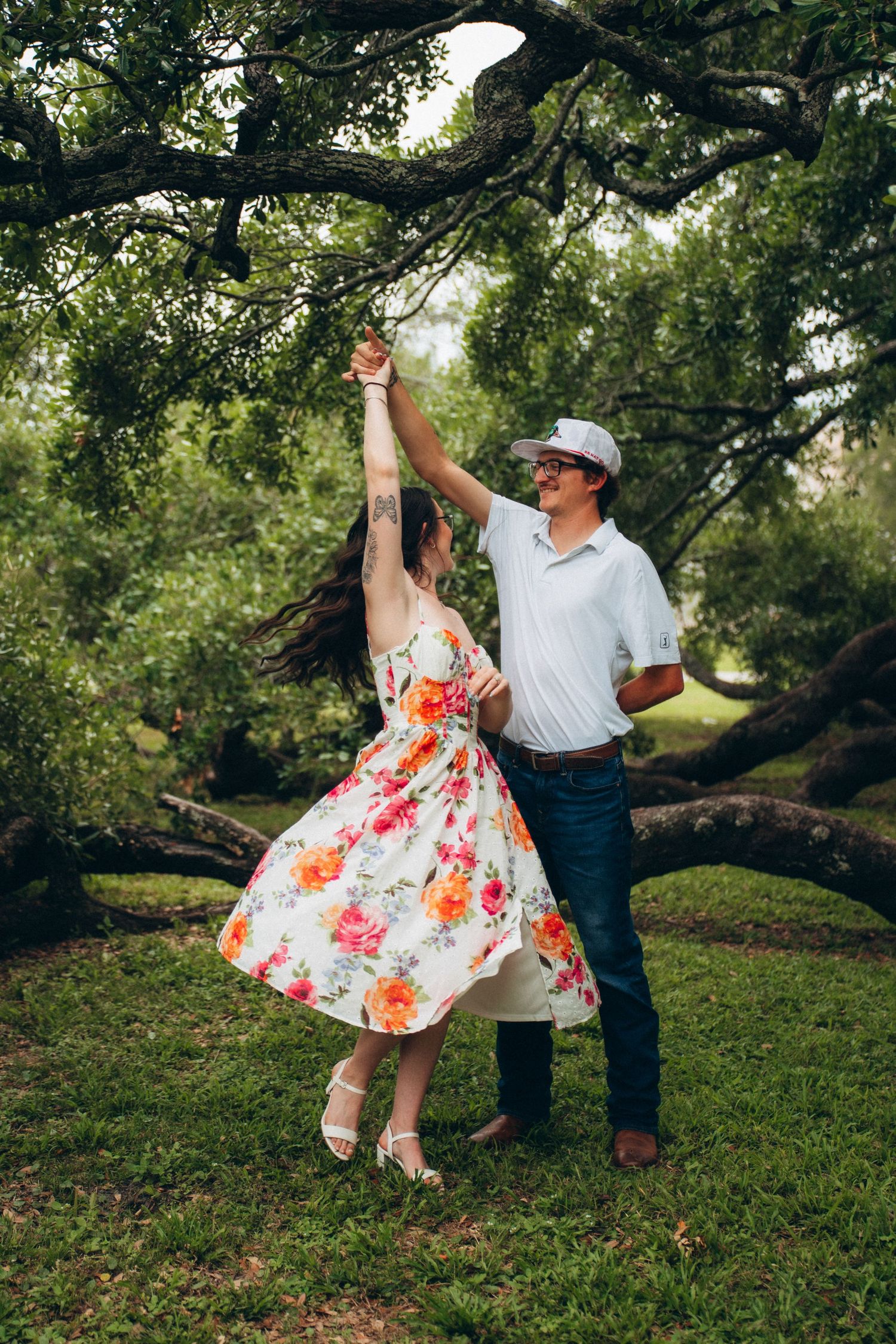 Couple dancing joyfully under oak trees, woman in floral dress being twirled by man in white shirt.