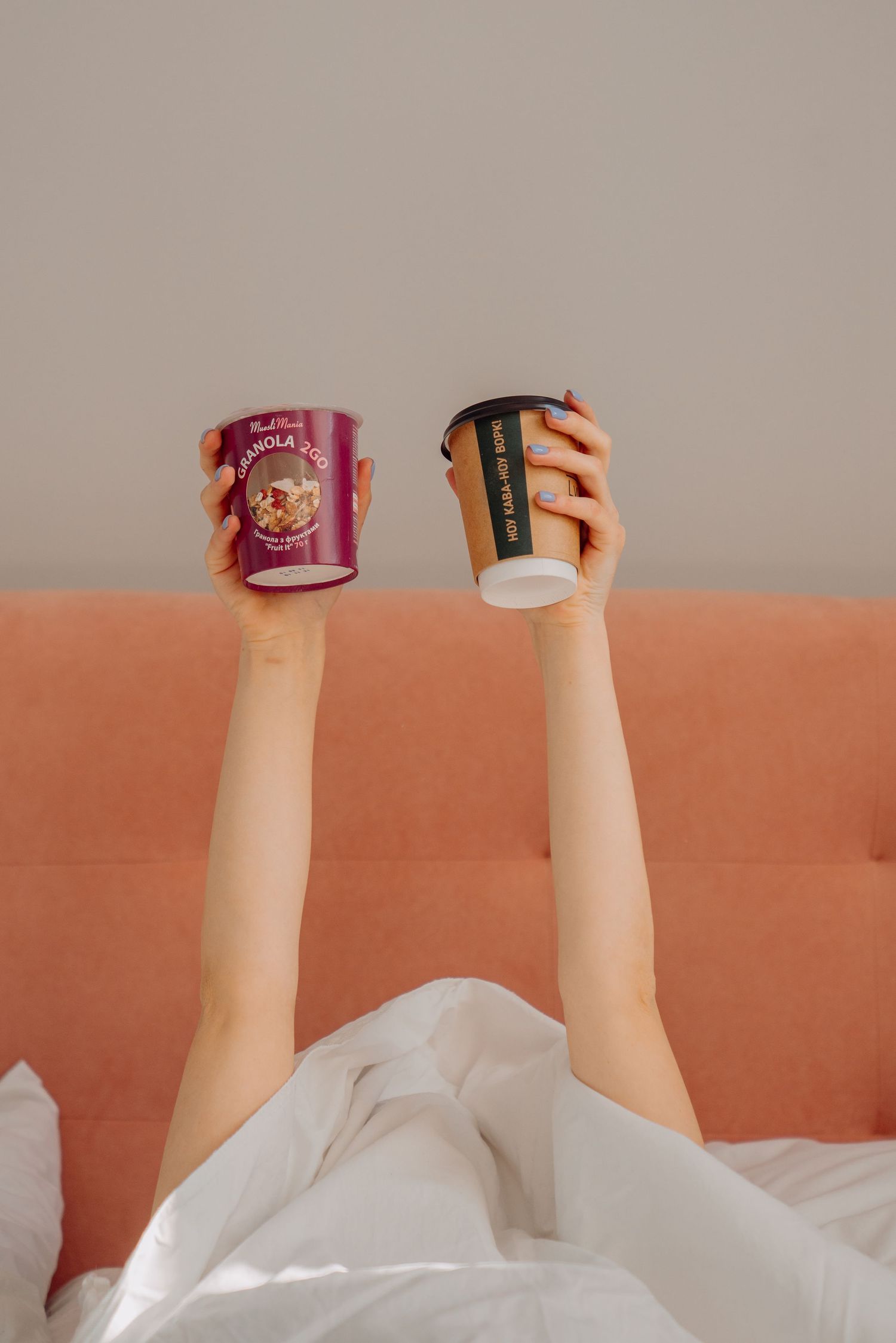 A person lying in bed holding up a granola cup and a coffee cup above a pink headboard.