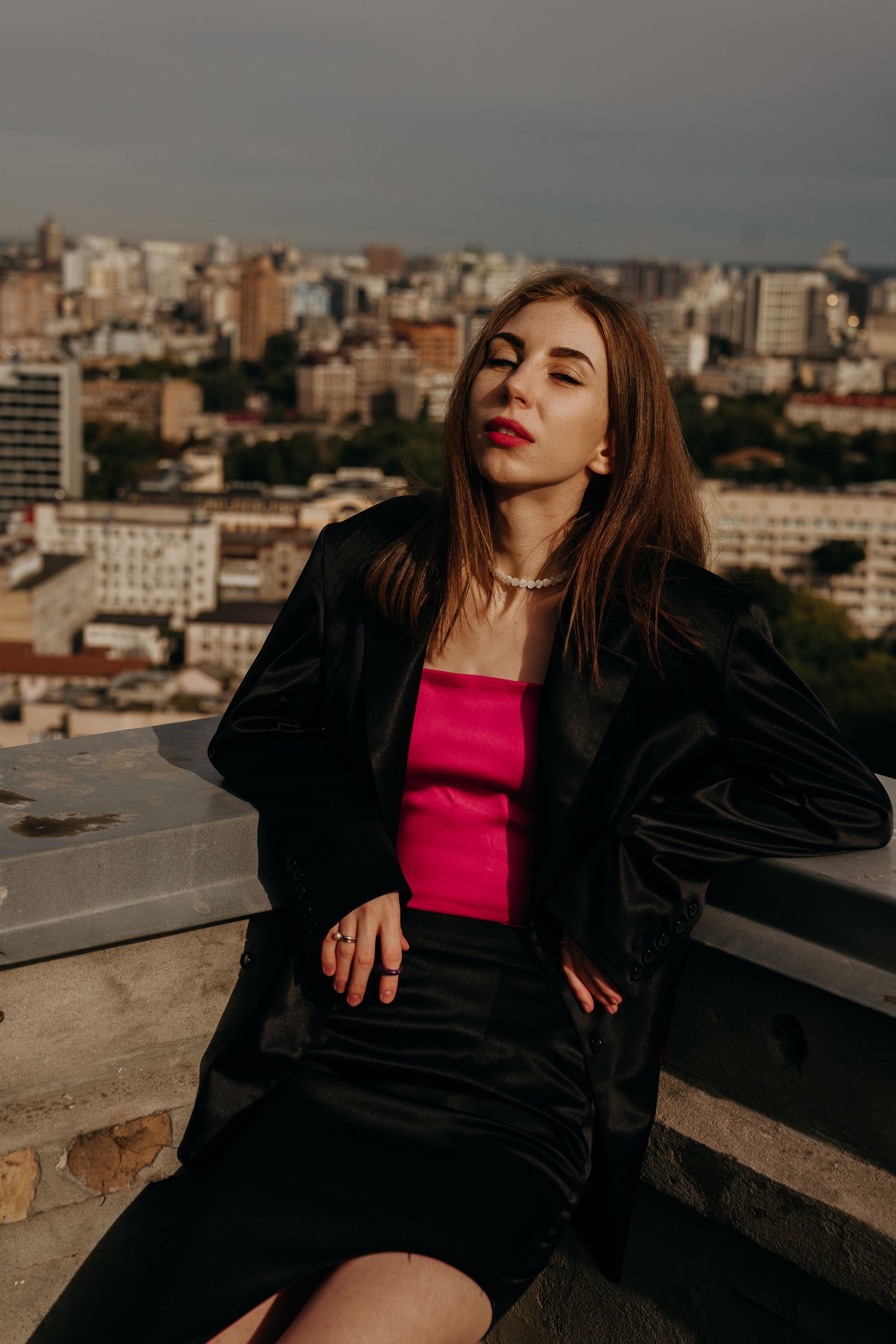 Young woman in black blazer and pink top poses on rooftop with city skyline backdrop.