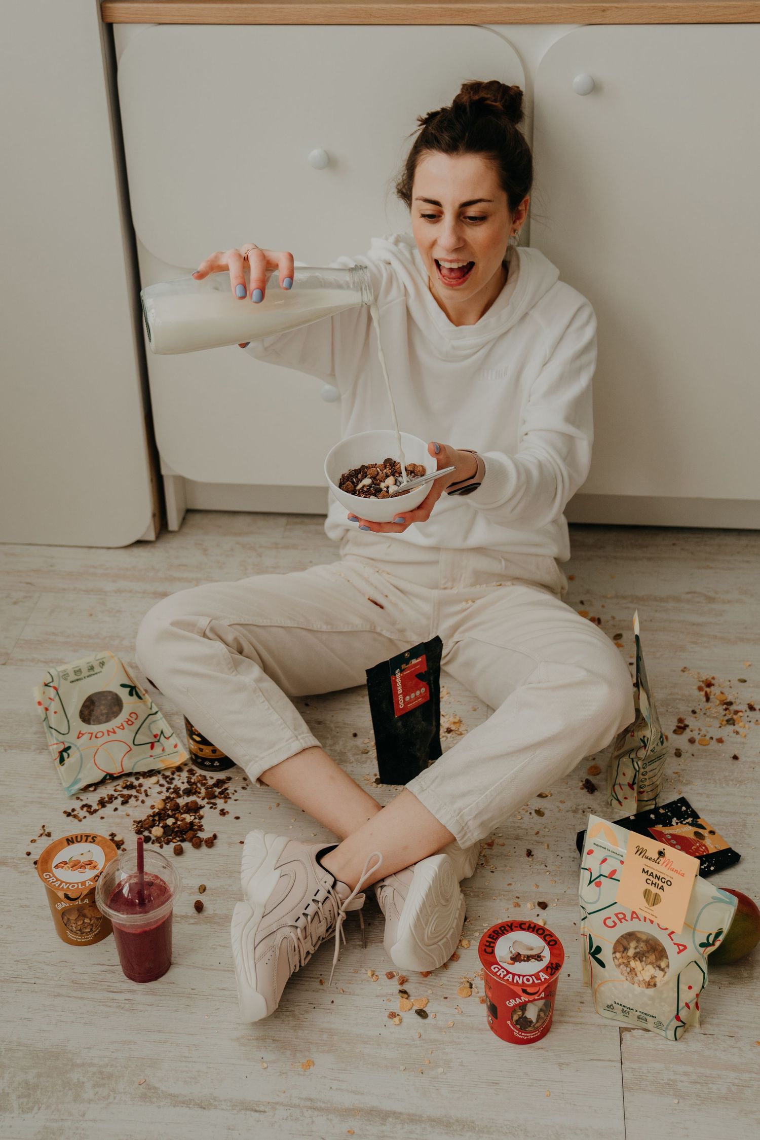 Woman in white outfit sitting on floor pouring milk into granola bowl surrounded by healthy snack products.