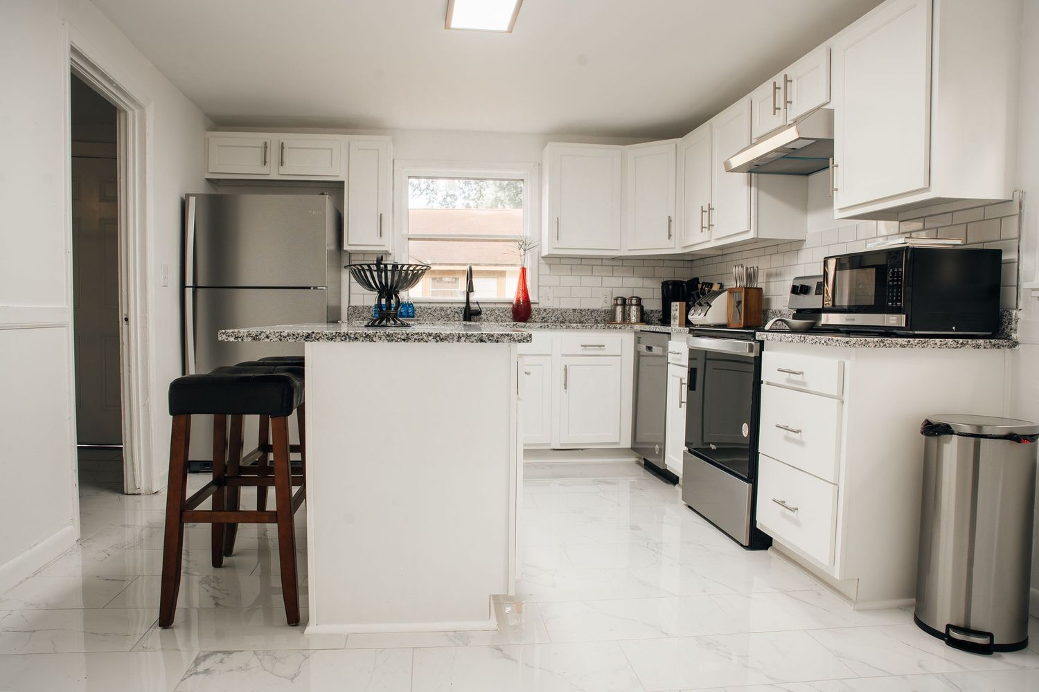 Modern white kitchen with stainless steel appliances, granite island, bar stool, and marble floors.