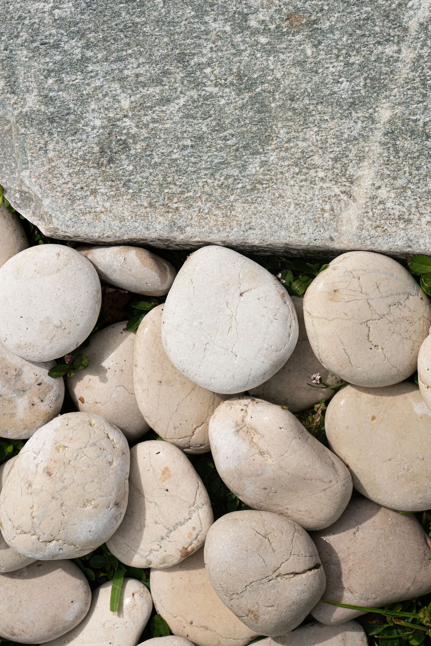 Close-up of rounded white cracked pebbles arranged beside a large flat grey granite stone on green grass.