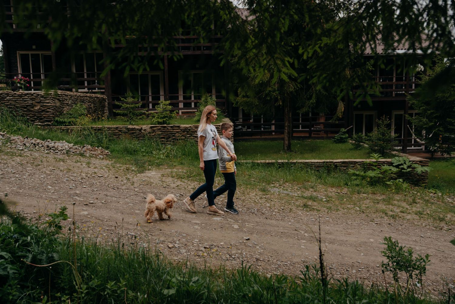 Two women walking a small golden dog on a dirt path near a rustic wooden building surrounded by lush green trees.
