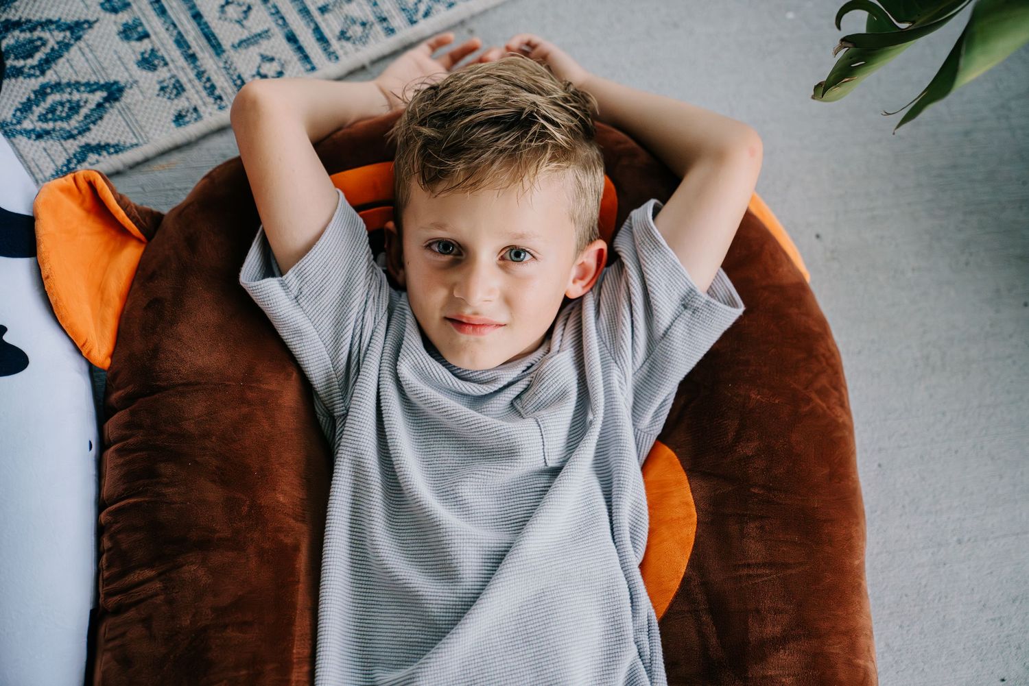 Young boy relaxing in a brown bean bag chair with hands behind head, wearing a gray t-shirt indoors.
