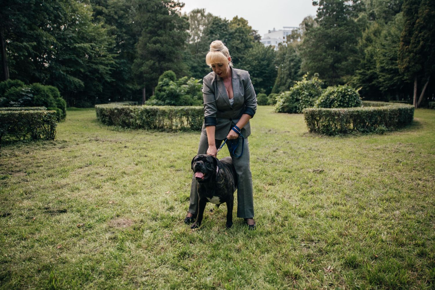 Blonde woman in gray suit bending down to pet a large black dog in a manicured park garden.