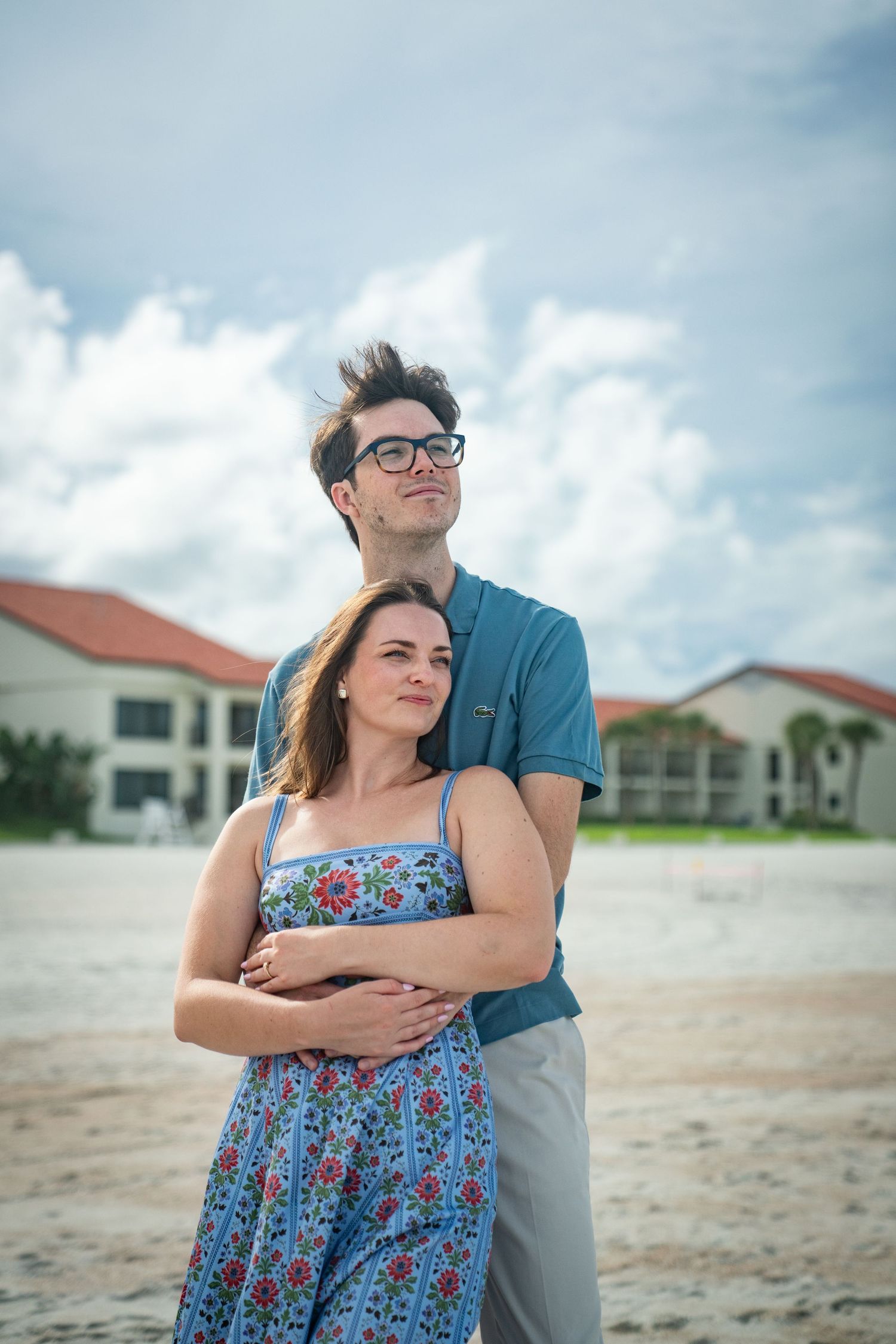 A couple shares a tender embrace on a sandy beach with white buildings in the background on a cloudy day.