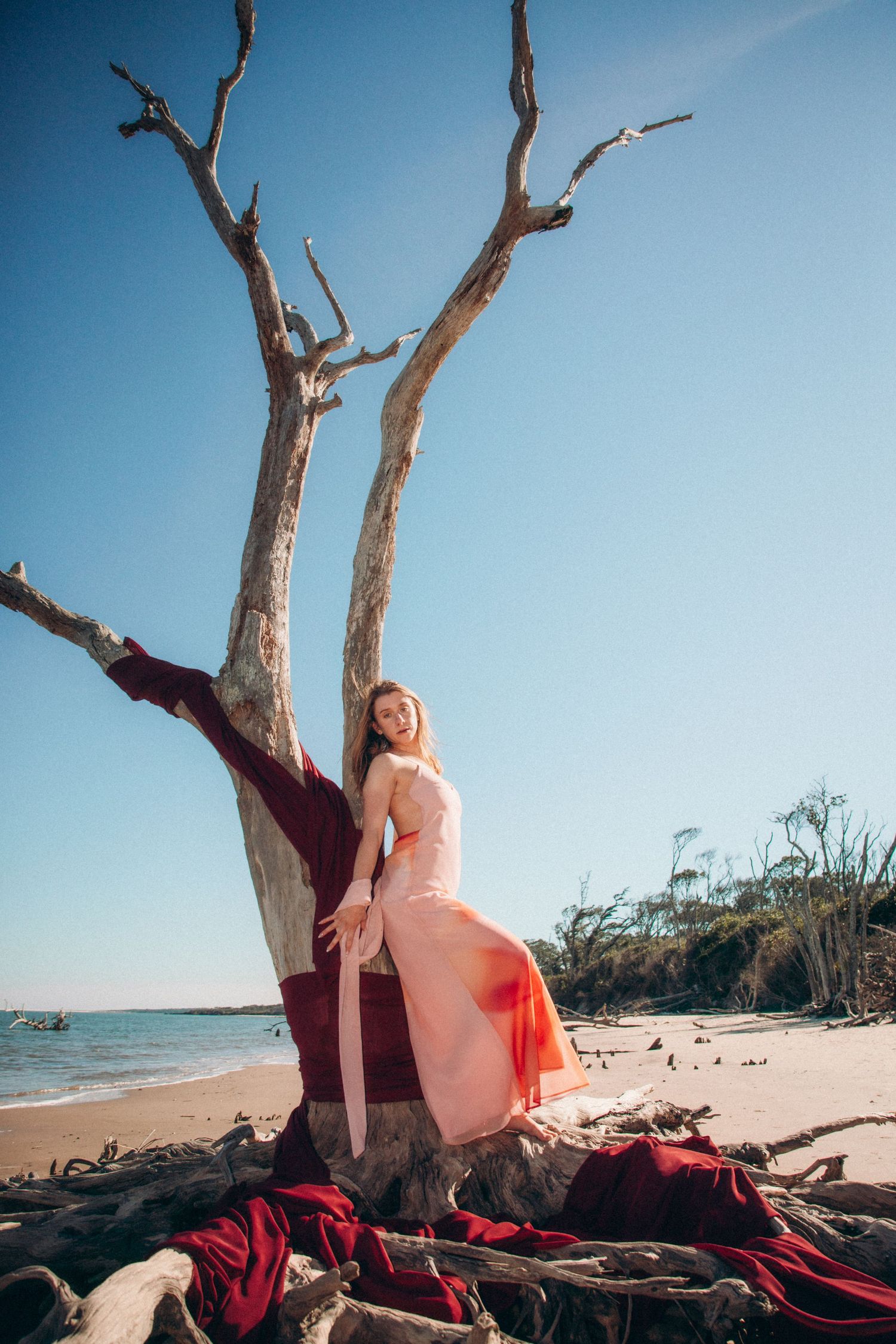Coastal photoshoot featuring driftwood and red fabric on a sunny beach with blue ocean views.