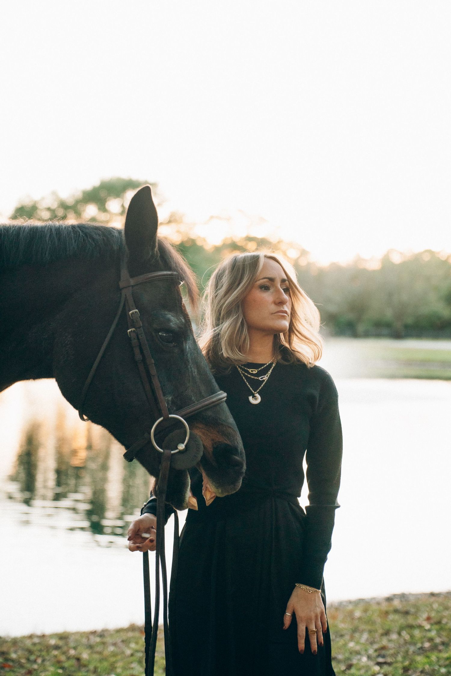 A woman in black stands beside a dark horse by a lake during sunset.