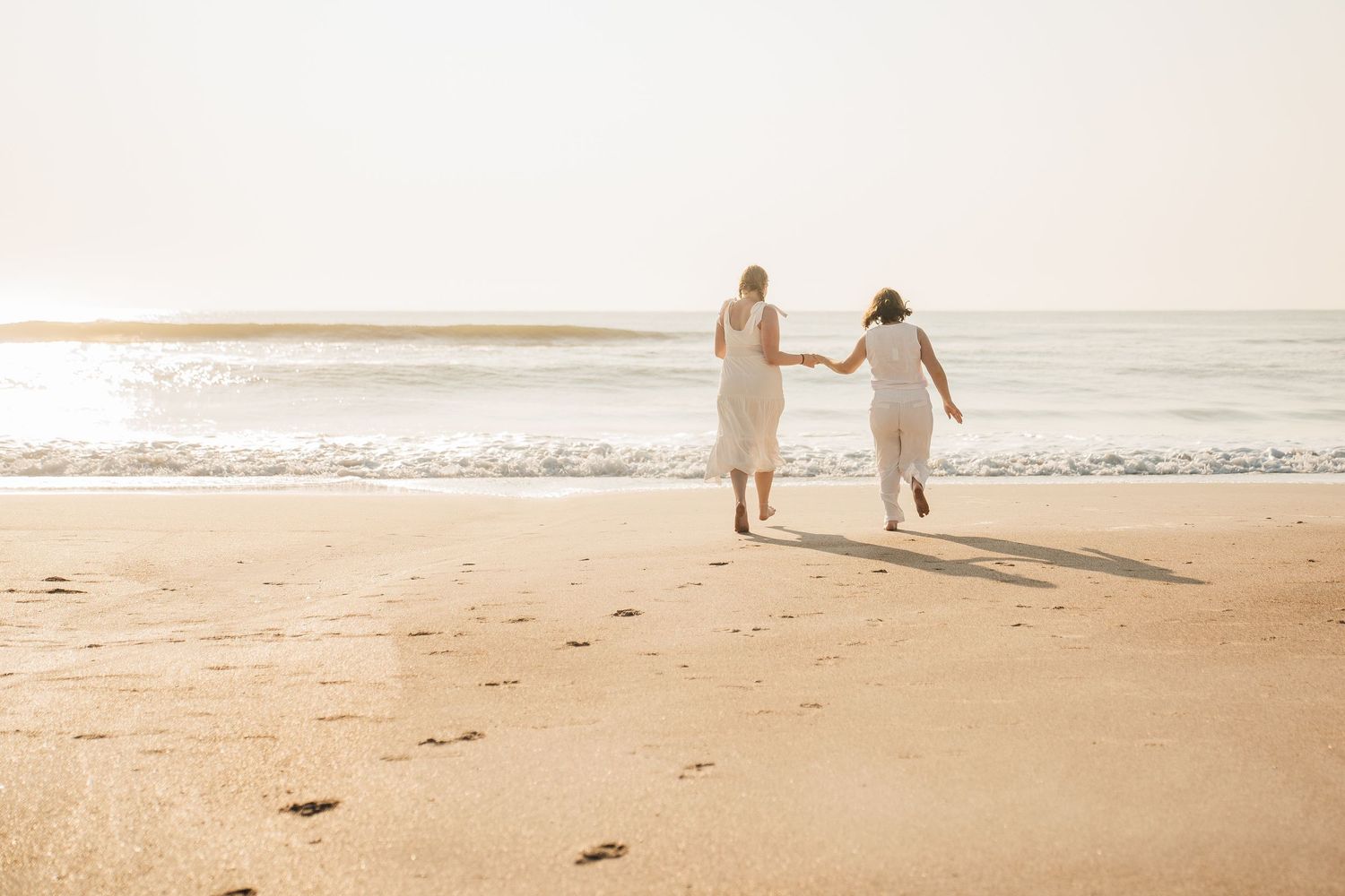 Two people in white dresses run along a sunlit beach with waves in the background.
