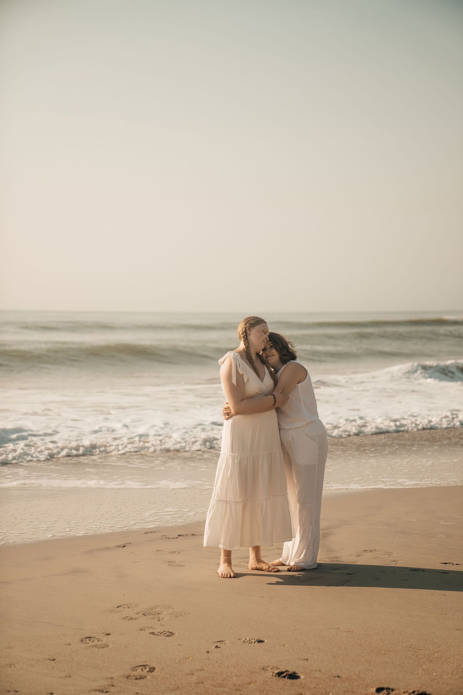 A couple in light-colored outfits embraces on a sandy beach with ocean waves in the background.