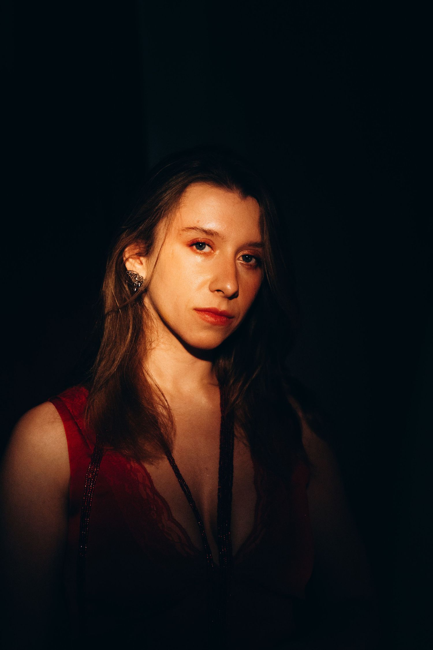 Young woman with long hair wearing a red top and earring, dramatically lit against a dark background.