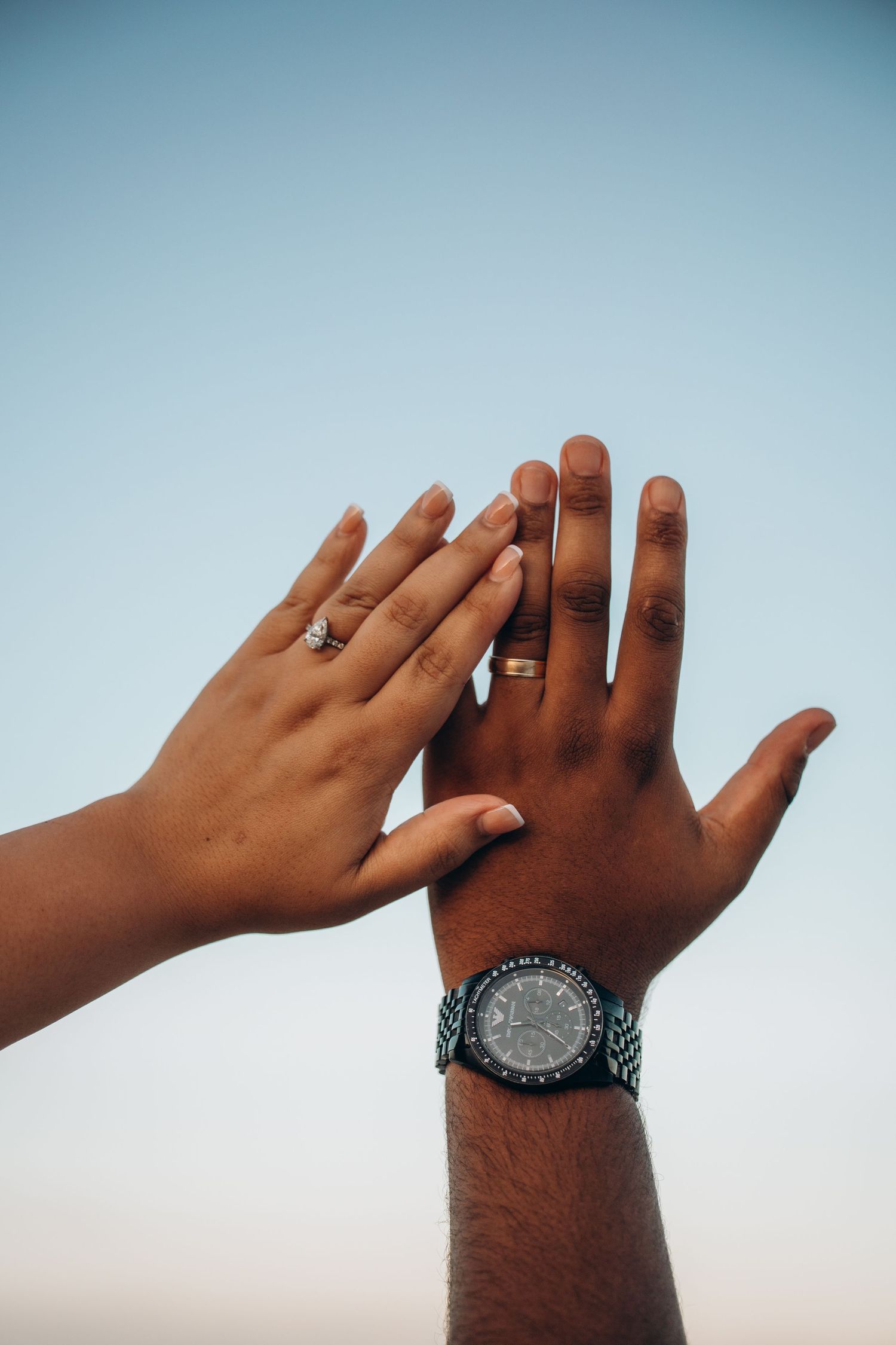 Couple raising hands together against blue sky, wearing wedding rings and a wristwatch, symbolizing unity.