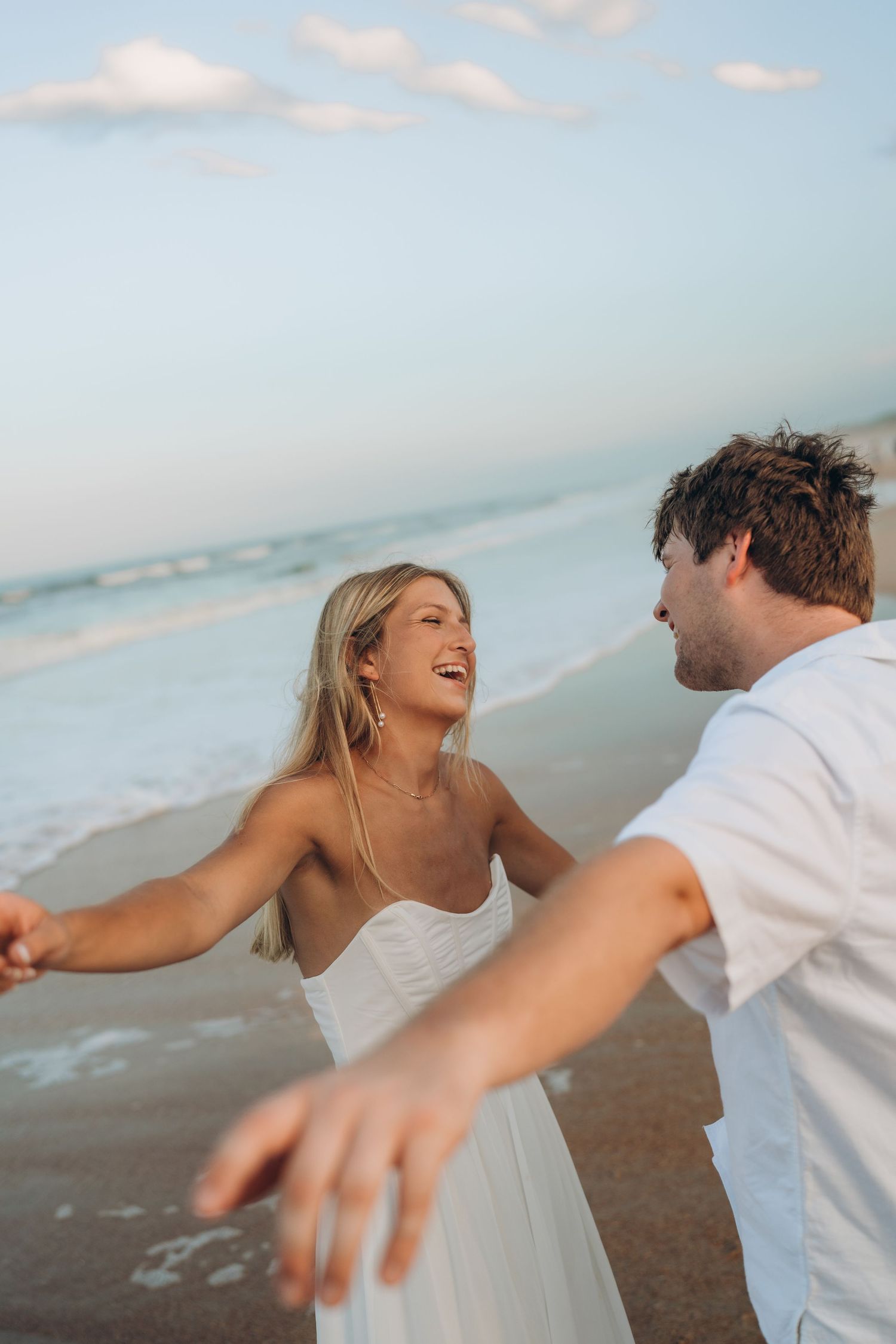 A couple in white outfits dances playfully on the beach at sunset with arms outstretched.
