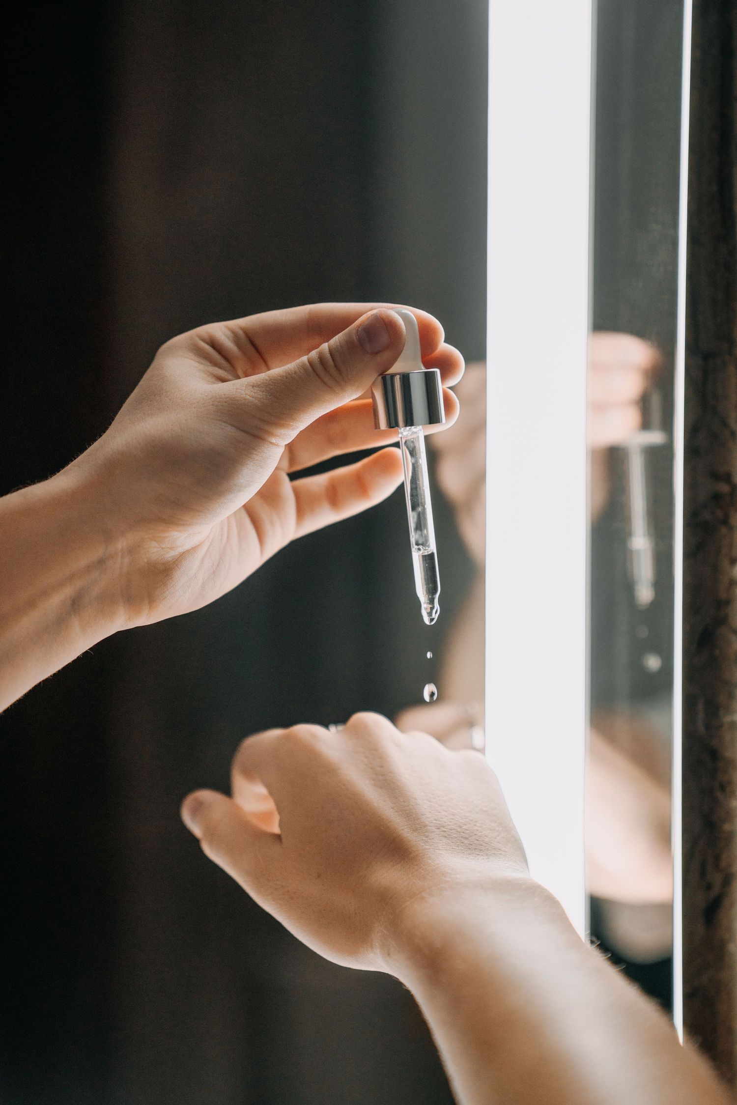 Close-up of a dropper dispensing clear serum drops near a lit mirror.