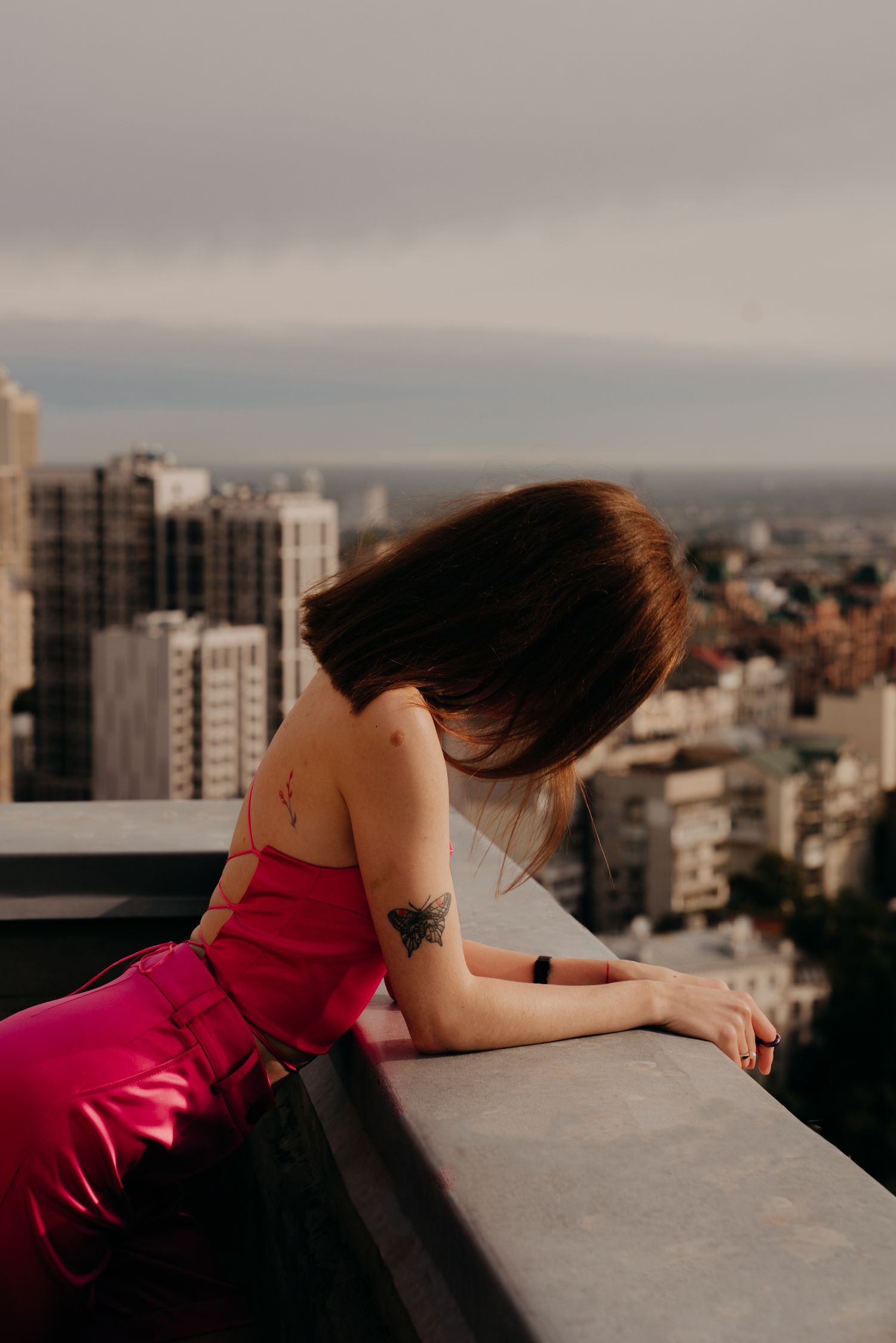Woman in pink dress with tattoos leans on rooftop railing overlooking city skyline on cloudy day.