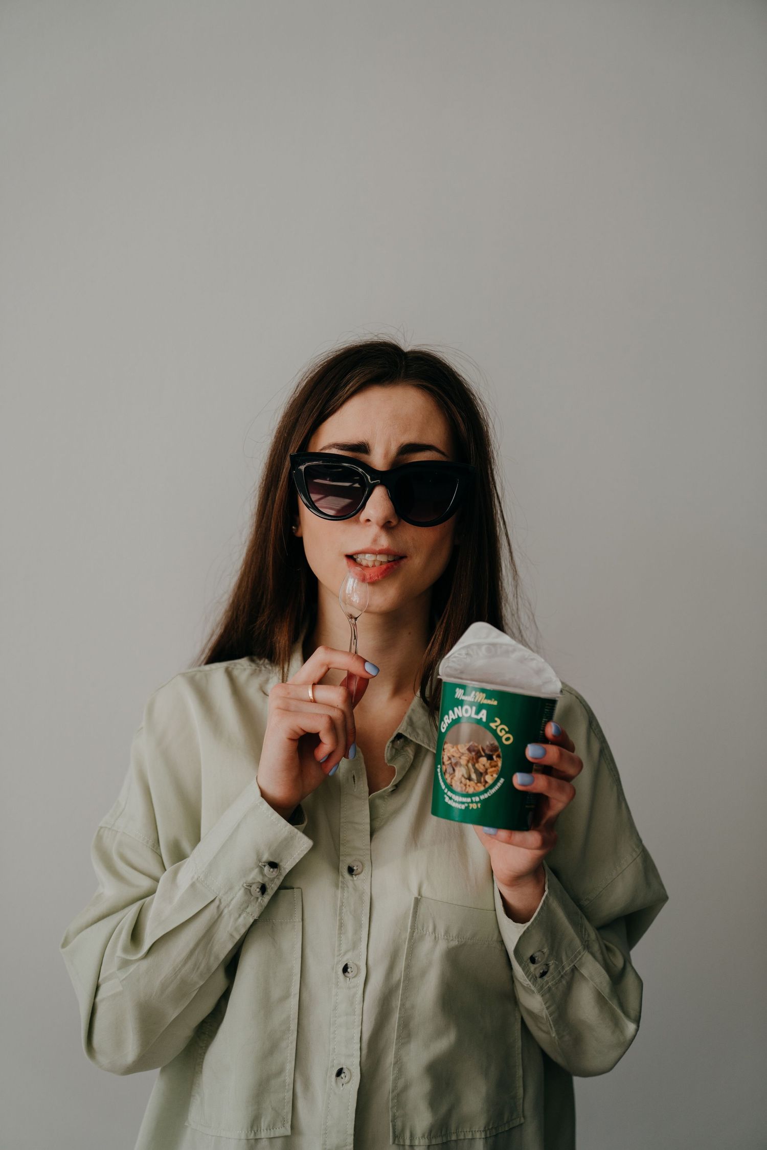 Young woman in sunglasses holding a green granola cup and spoon, wearing a sage button-up shirt against a gray background.
