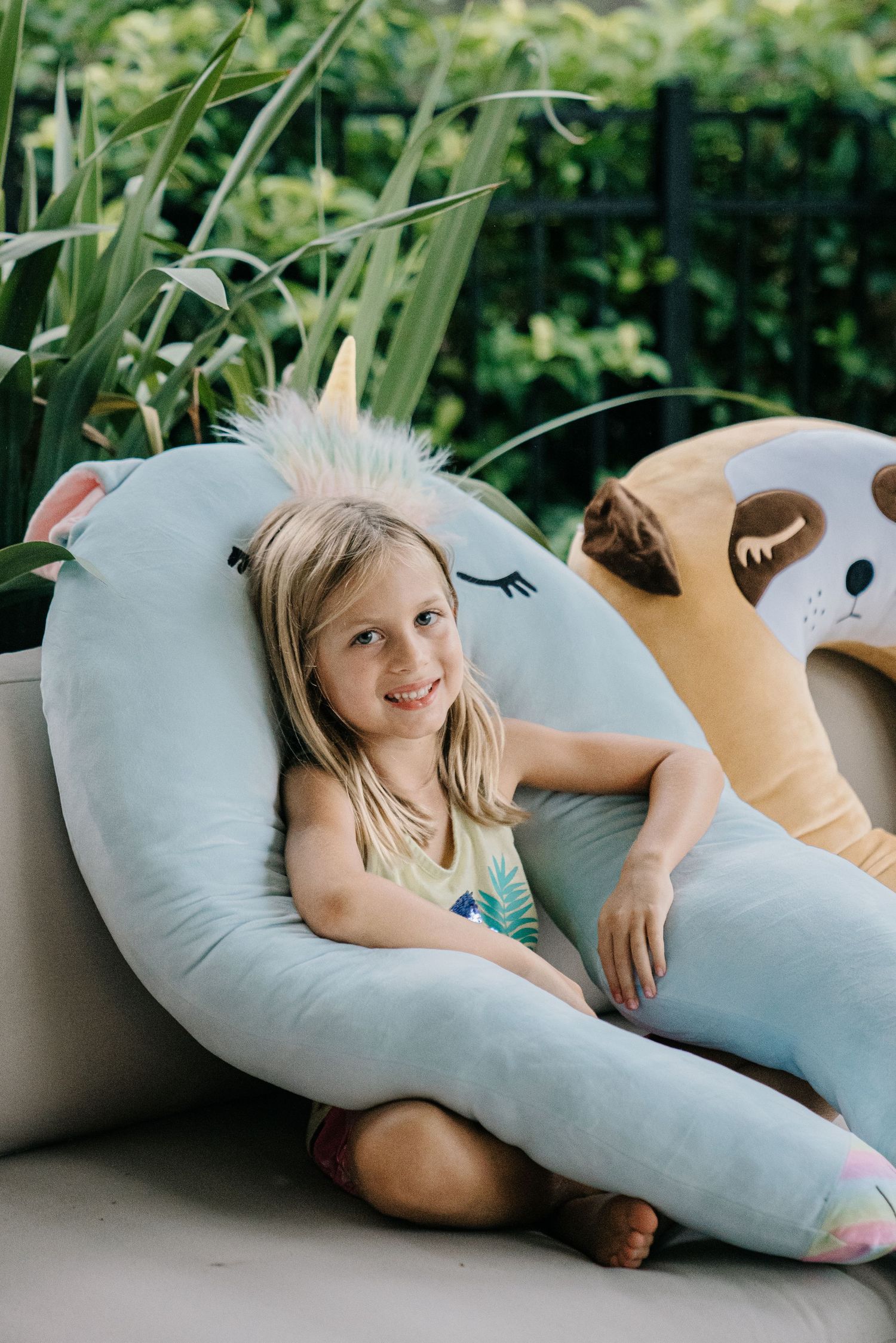 Young girl smiling while sitting in a unicorn-shaped plush chair outdoors surrounded by green plants.