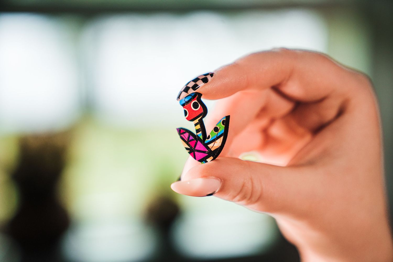 Hand holding a small colorful decorative anchor pin with vibrant multicolor patterns against a blurred background.