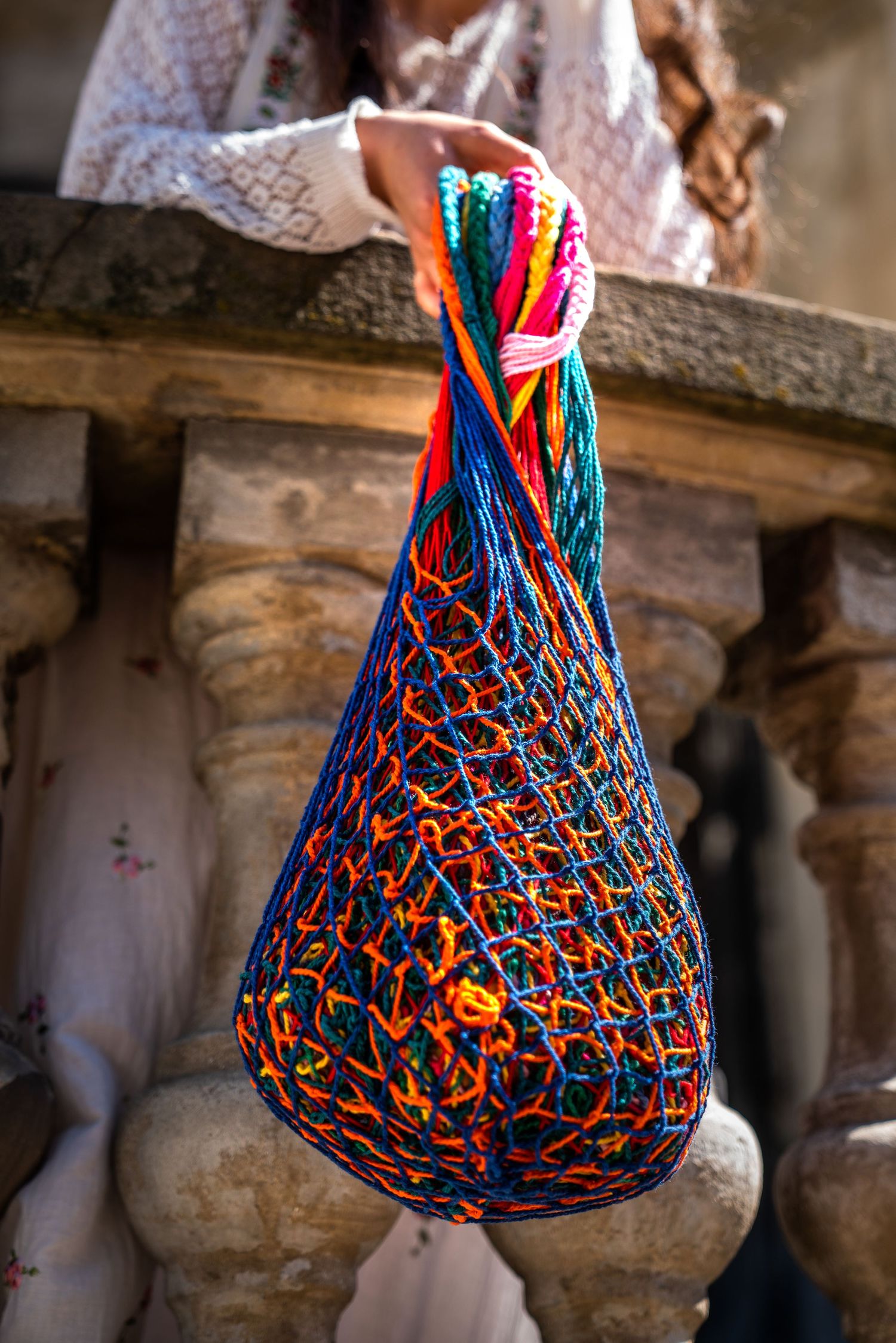 Woman holding a colorful blue and orange handmade crochet net bag outdoors near stone balustrade.