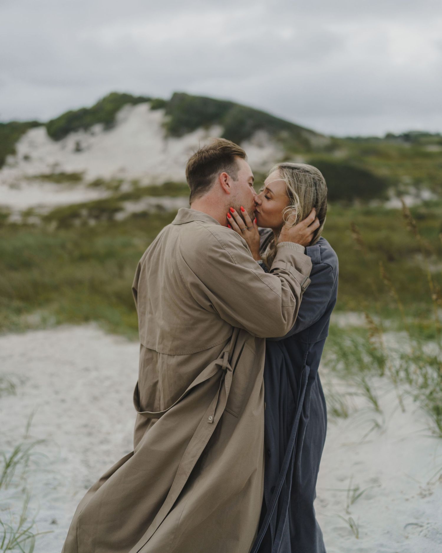 A romantic couple sharing a tender moment on a sandy path with green hills and cloudy skies in the background.
