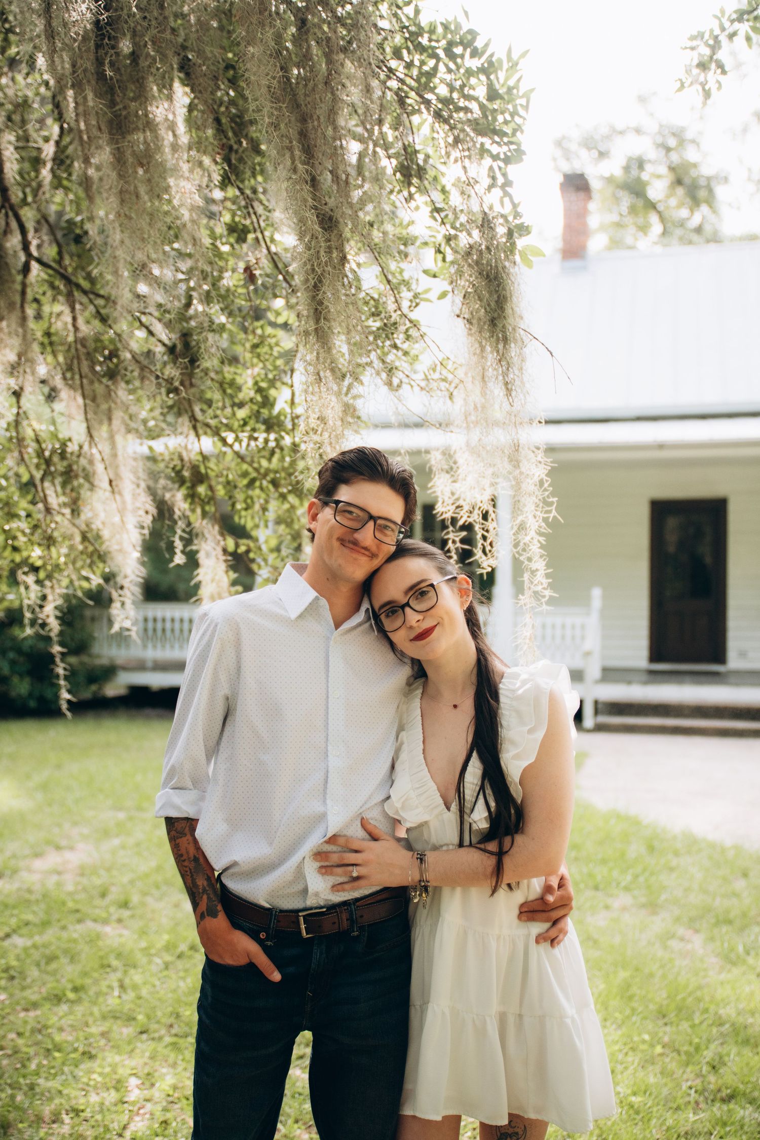 A couple wearing glasses embrace under Spanish moss hanging from trees in front of a white farmhouse.