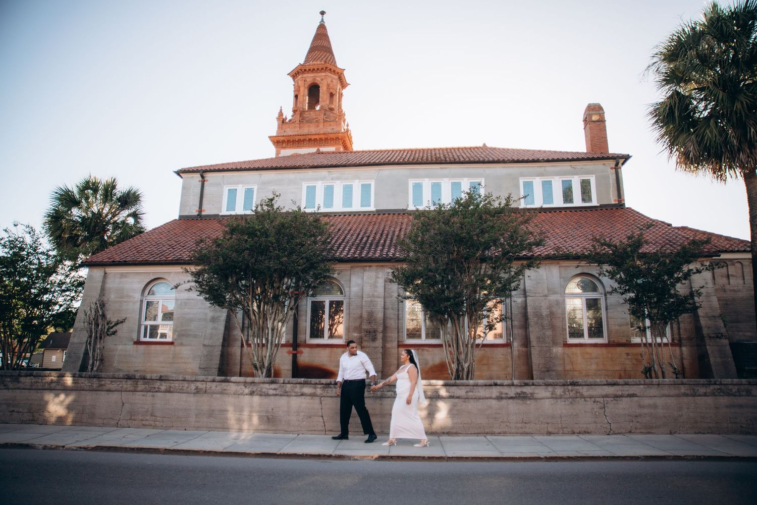 Bride and groom hold hands outside a historic stone church with a red tile roof and bell tower at dusk.