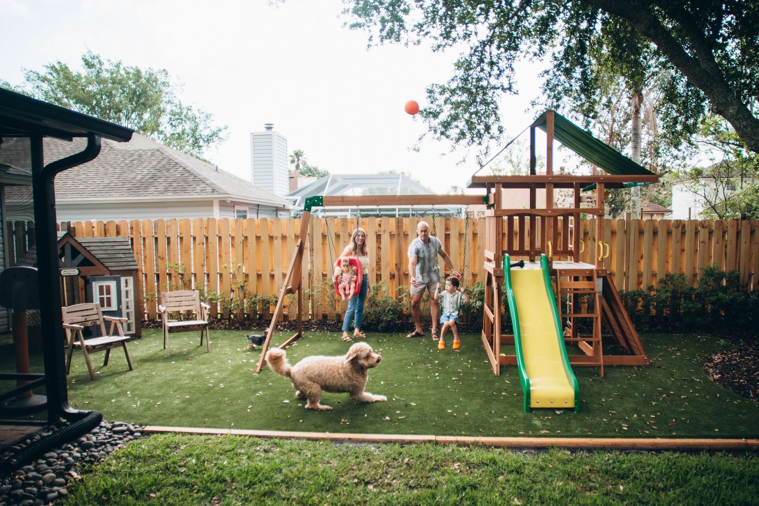 Family with children and golden dog enjoying backyard playset with green slide and swing set on artificial turf.