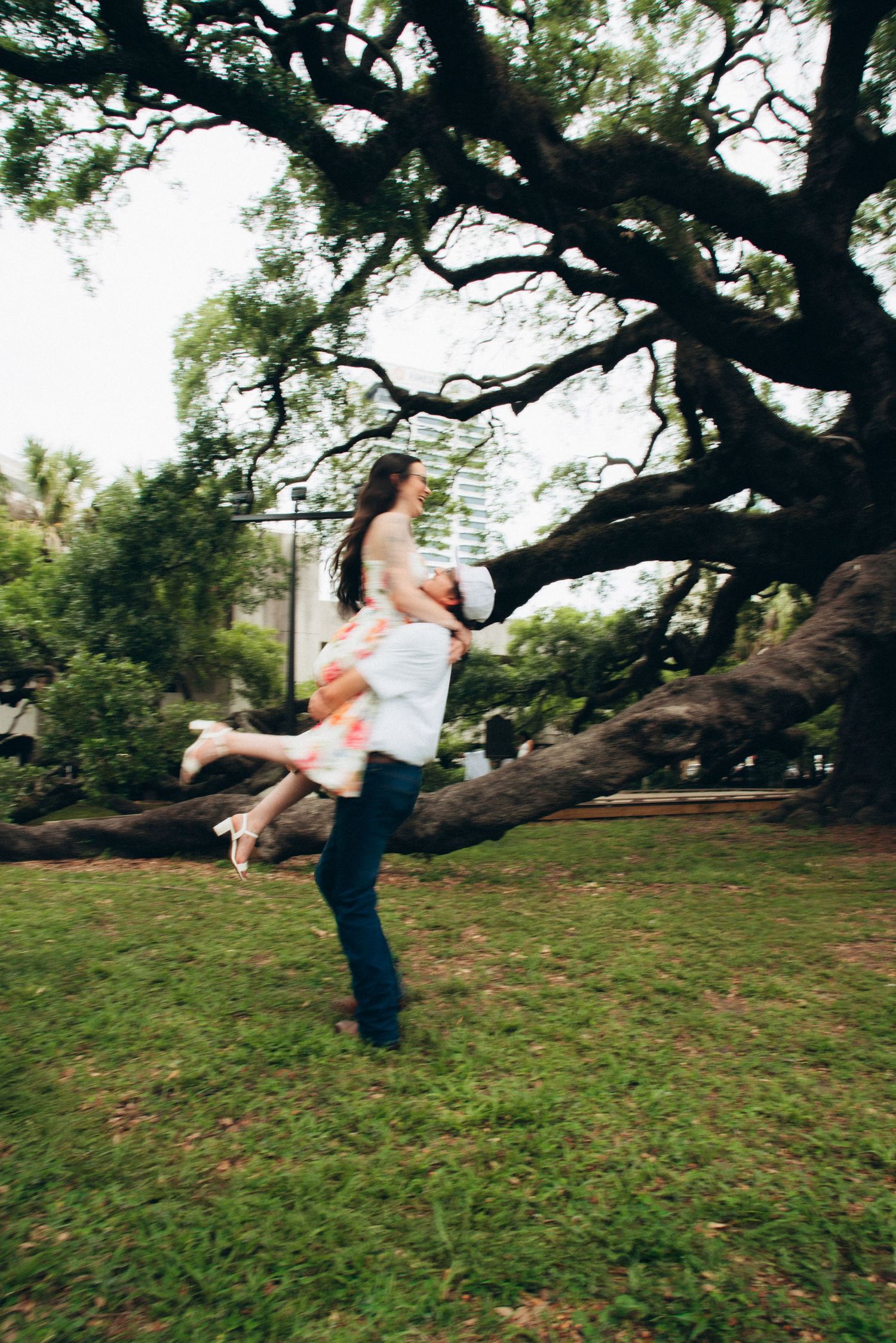 Man lifting woman in floral dress outdoors under large tree during romantic engagement photo session.
