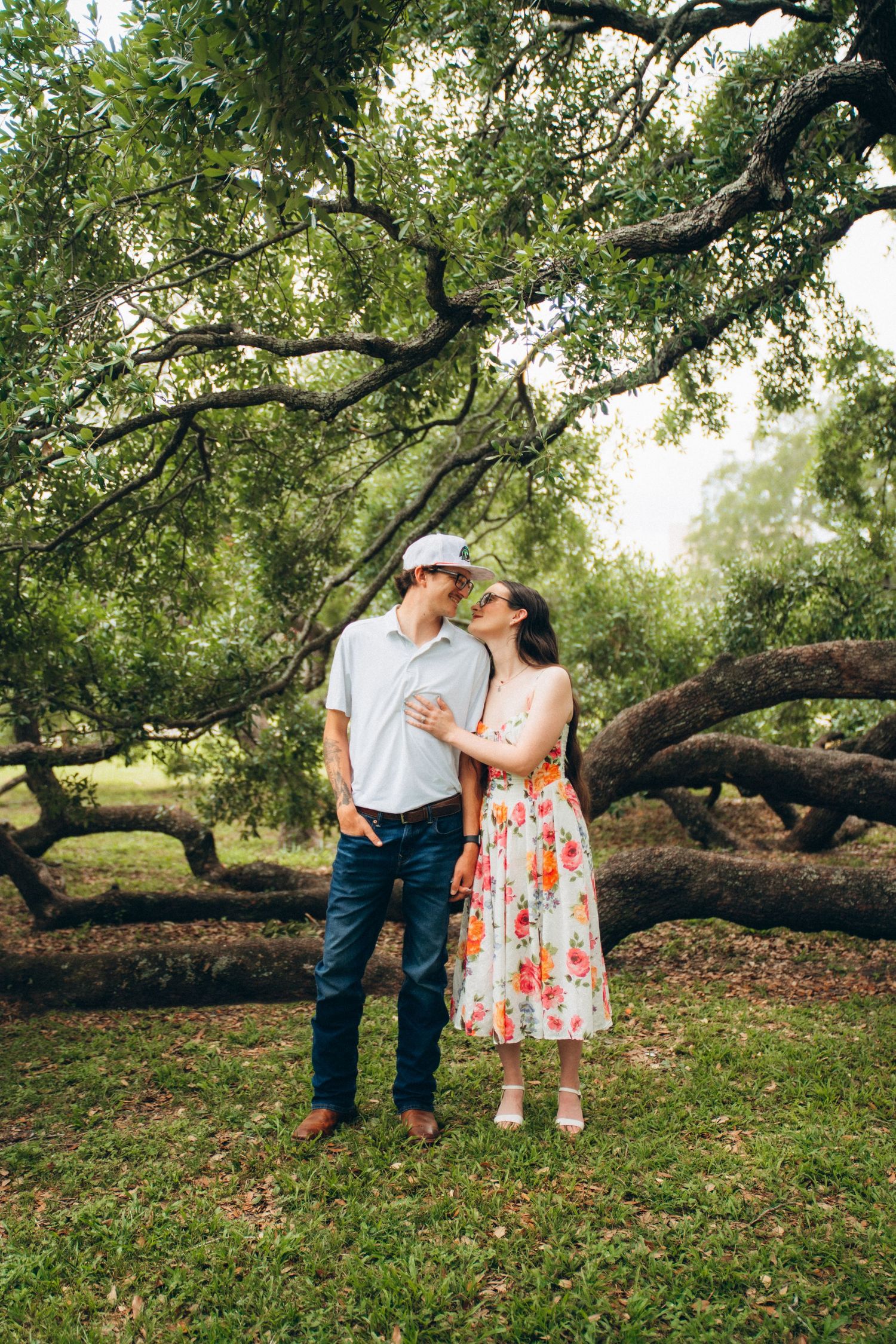 Couple sharing a tender moment under sprawling oak trees, woman in floral dress kissing mans cheek.