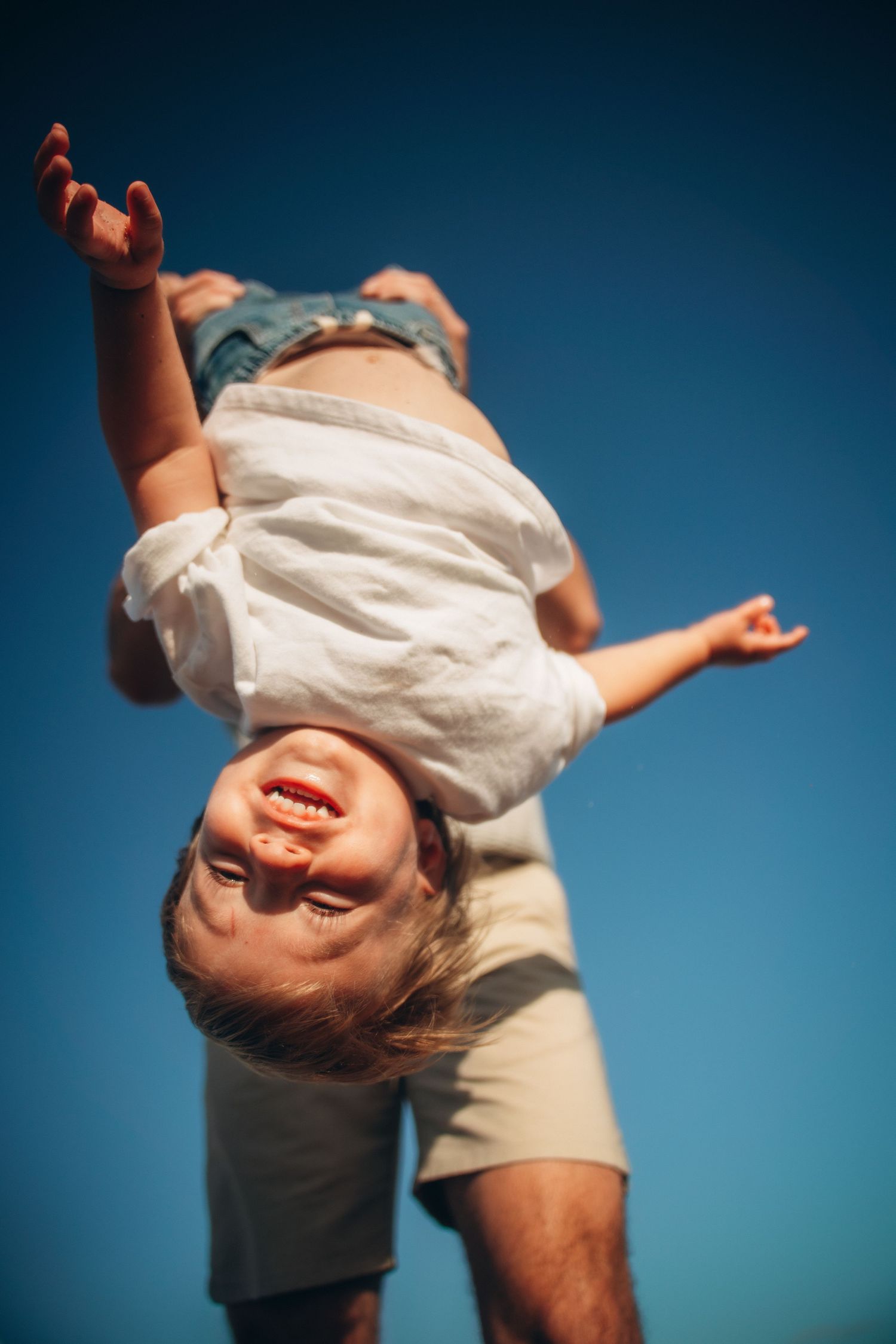 A joyful toddler held upside down by a parent against a clear blue sky, laughing with arms outstretched.