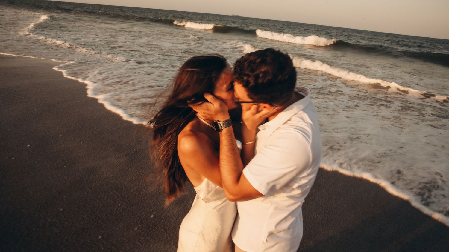 A romantic couple shares a passionate kiss on a sandy beach at sunset with ocean waves in the background.