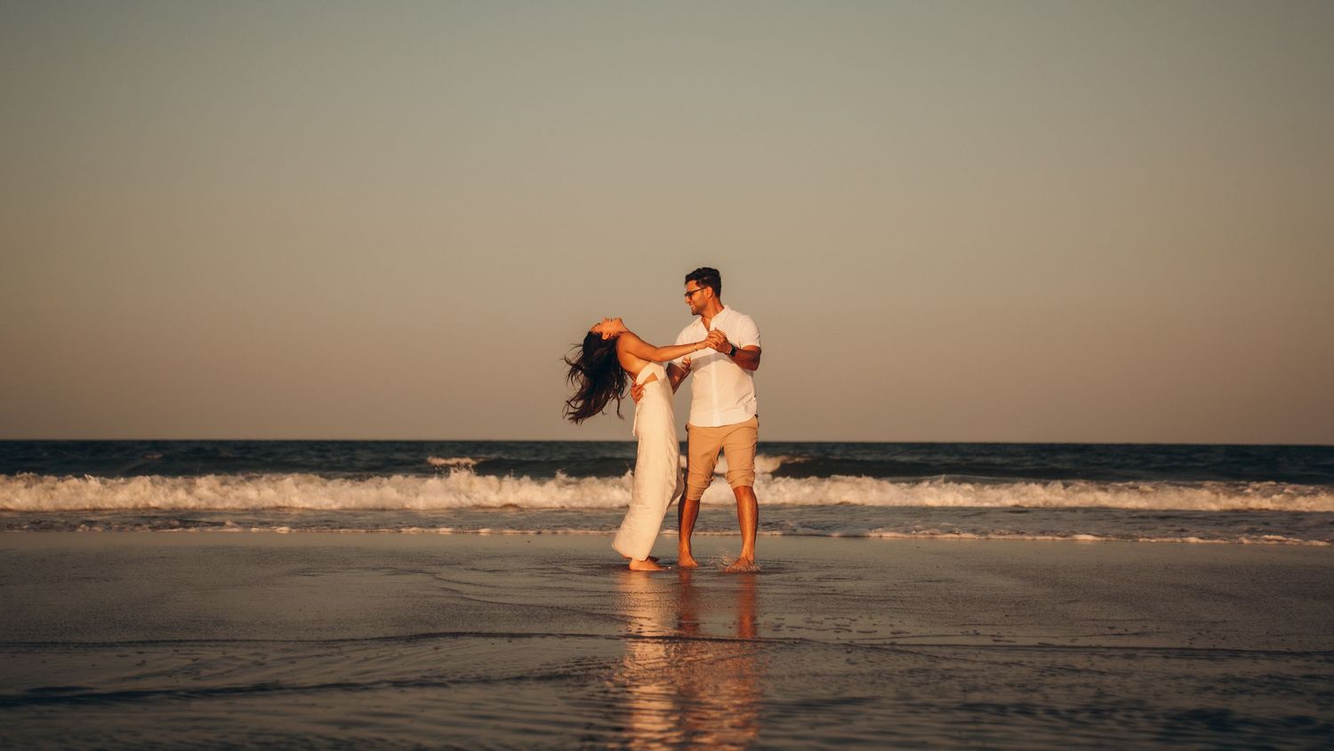 A romantic couple dancing together on a sandy beach at sunset, waves gently washing around their feet.