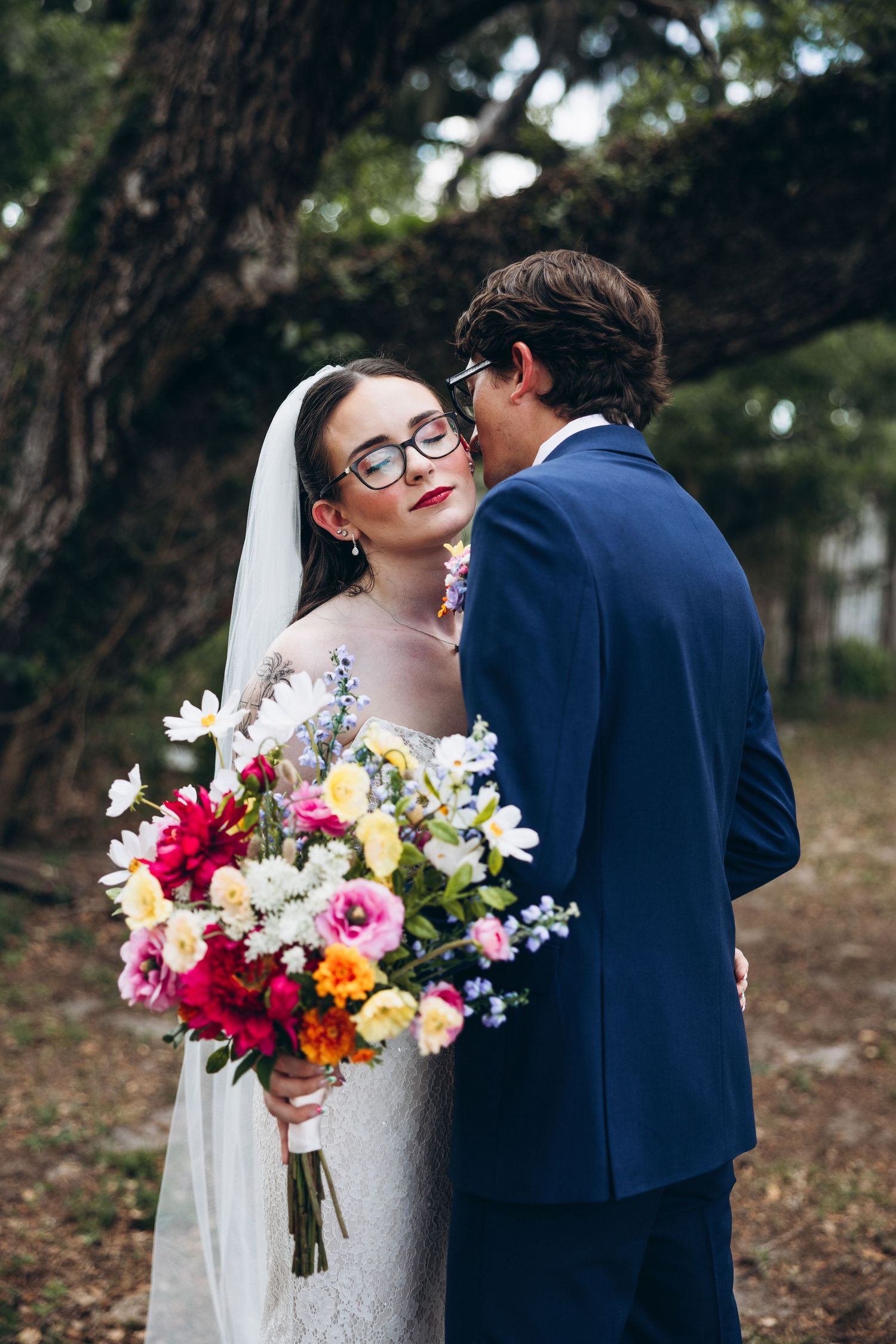 Bride in lace gown with colorful bouquet and groom in navy suit share a tender moment under trees.
