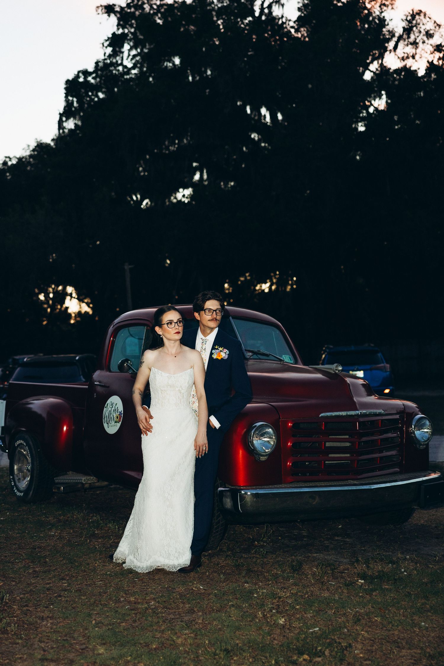 Bride and groom in wedding attire posing in front of a vintage red truck at dusk with trees in background.