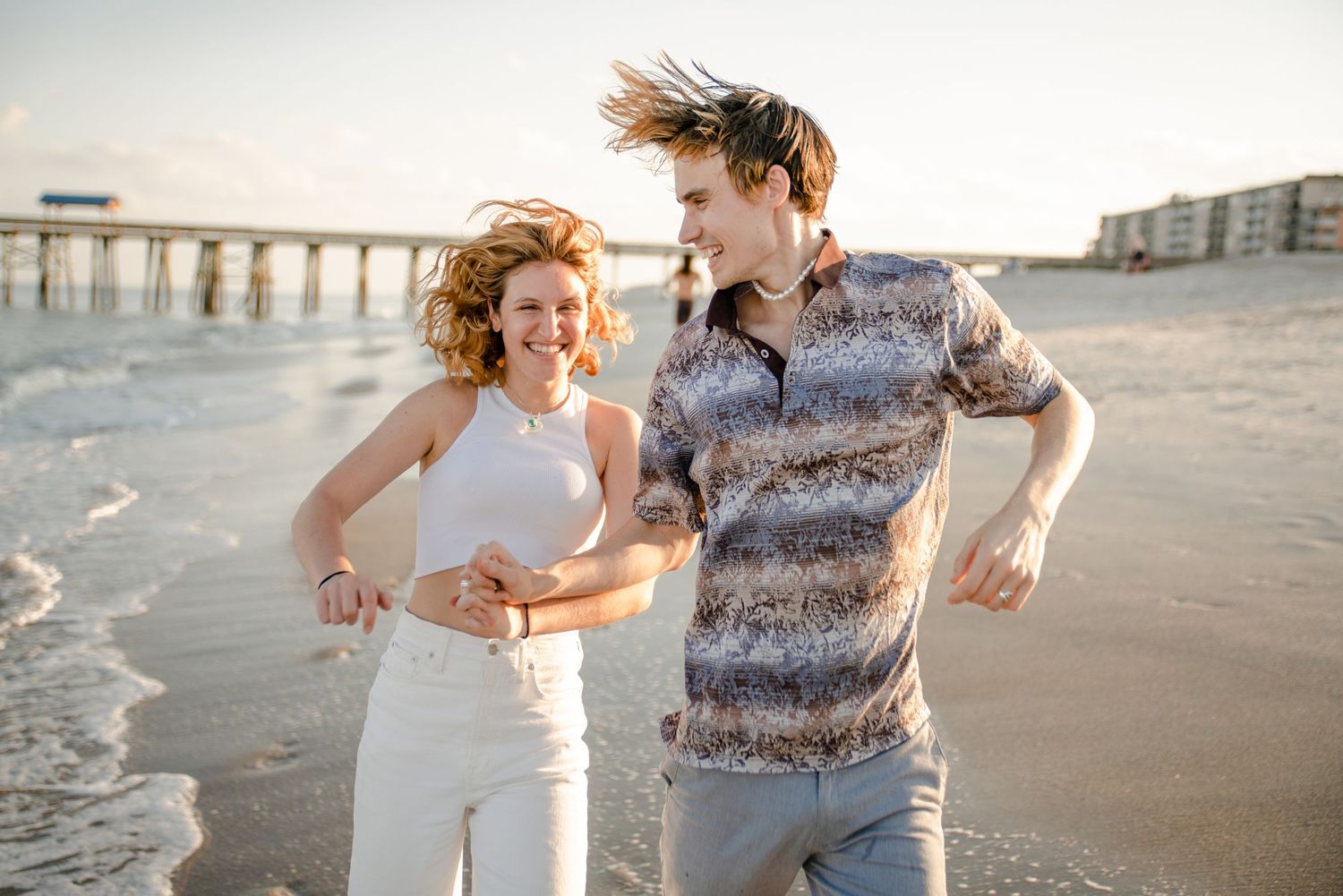 Joyful couple walking along beach with ocean waves and buildings in background.