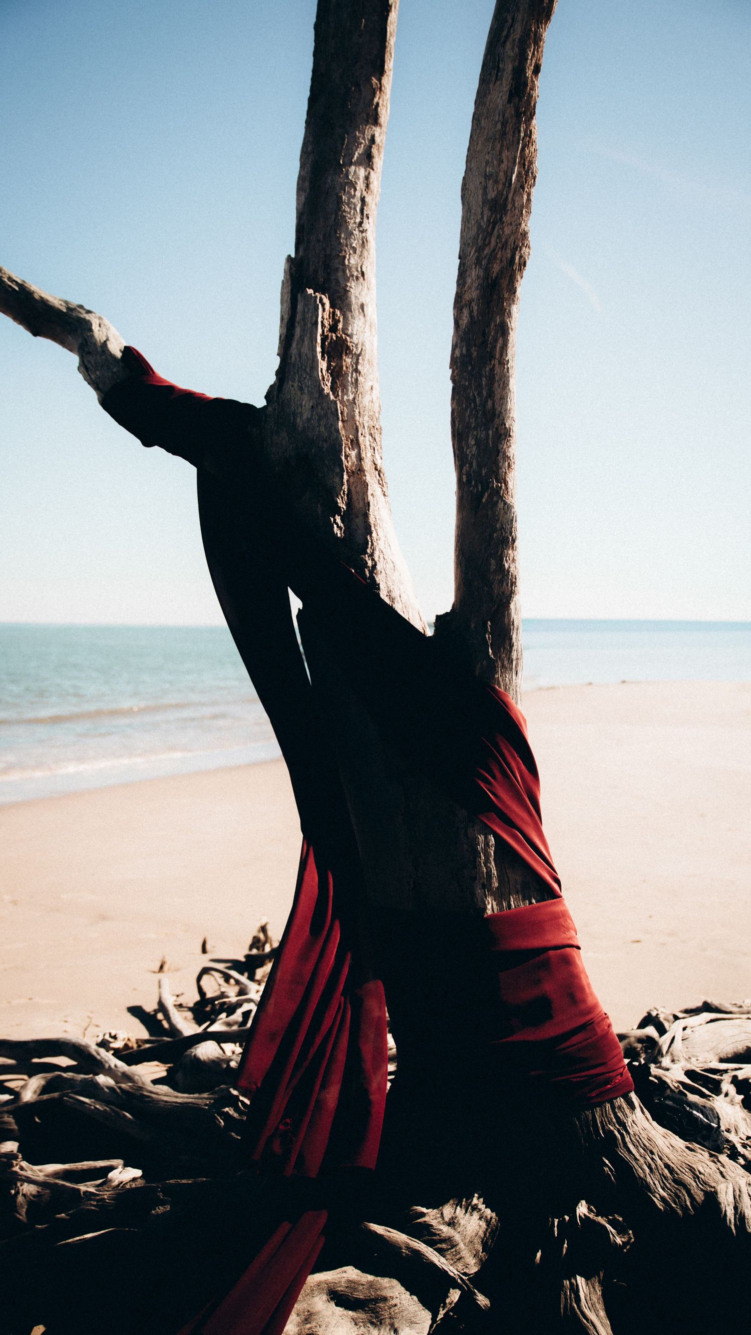 Red fabric wrapped around a weathered tree trunk on a sandy beach with ocean view.