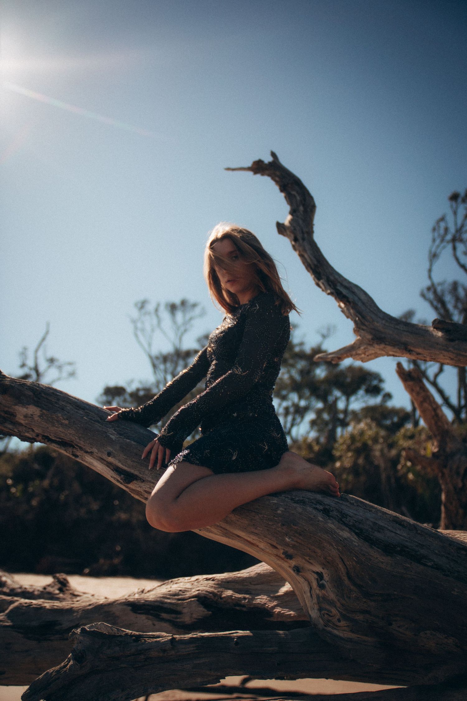 A figure in dark clothing poses gracefully on large driftwood pieces at the beach.