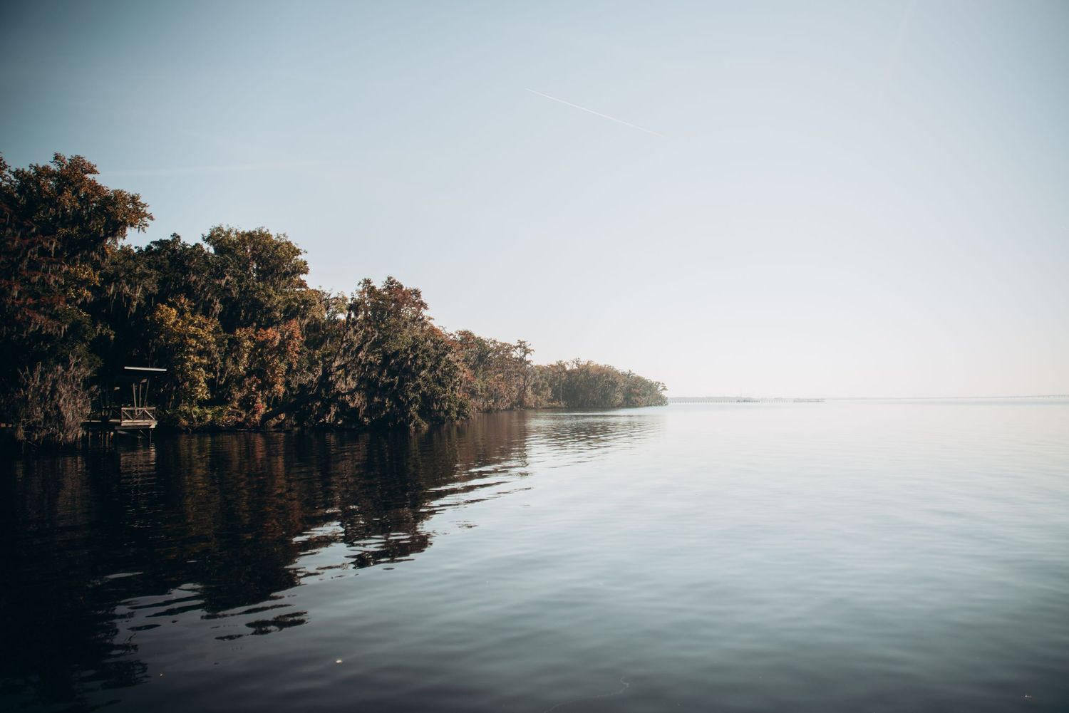 Calm lake waters reflect autumn trees along a misty shoreline during a serene morning.
