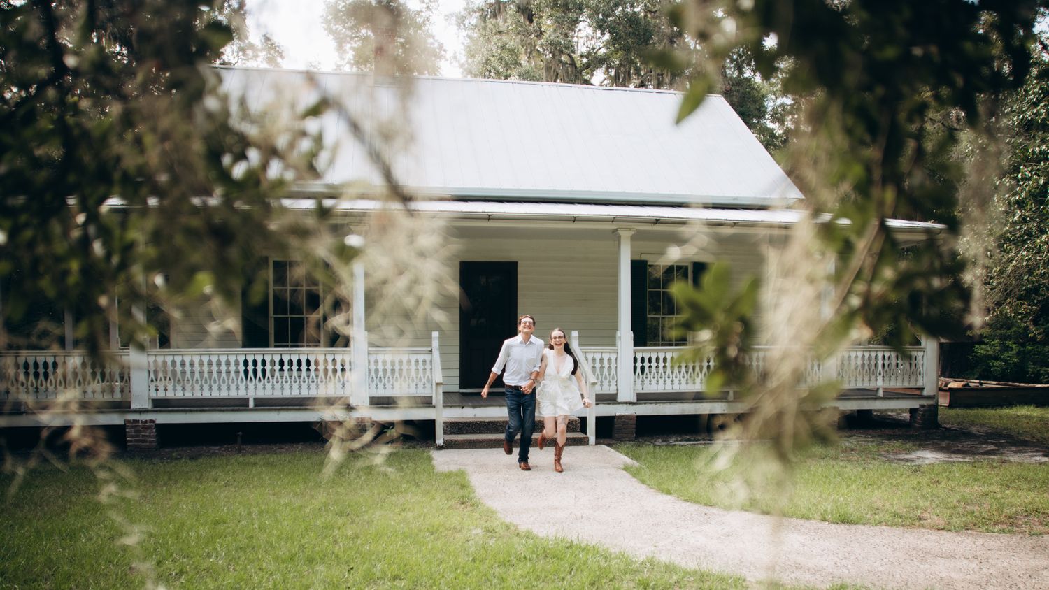 Couple walking hand in hand on a path in front of a charming white Southern farmhouse with a wraparound porch.